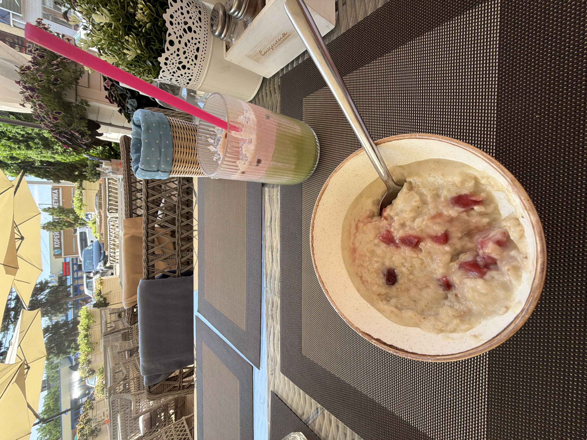A close-up, eye-level shot captures a breakfast scene on an outdoor patio, likely in Leningradskaya, Russia. In the foreground, a bowl of oatmeal with visible red berry pieces sits on a dark brown placemat. A silver spoon rests inside the bowl, partially submerged in the oatmeal. To the right, a tall glass contains a layered beverage, appearing to be a smoothie or juice, with a bright pink straw. Behind the drink, a white decorative pot holds a lush green plant. Further back, a wicker basket with a blue polka-dot lining is visible. The background reveals a bustling outdoor seating area with rattan chairs and tables, some covered with dark fabric. Beyond the patio, a street scene unfolds with buildings, parked cars, and pedestrians under a bright, sunny sky, suggesting a daytime setting. The overall impression is a pleasant outdoor meal experience.