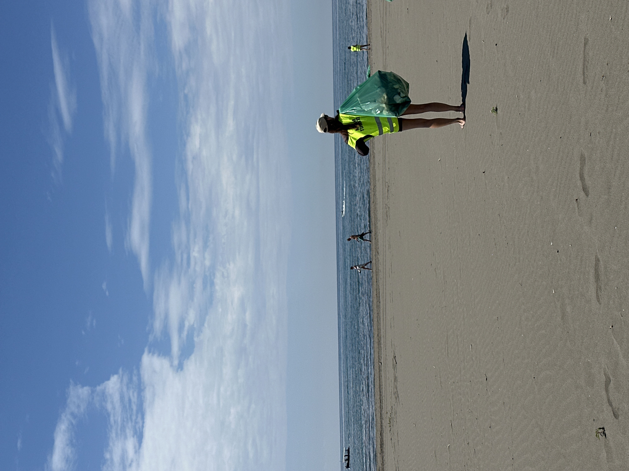 The image shows a person wearing a yellow vest and a cap standing on a sandy beach in Donji Štoj, Montenegro. The person is holding a green plastic bag. The beach is wide and flat, and the sea is visible in the background. There are other people walking along the beach in the distance. The sky is blue with some white clouds. There are some footprints on the sand.