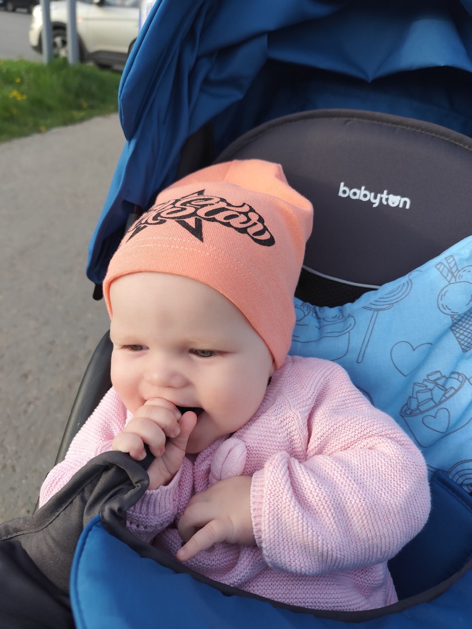 A baby sits in a blue stroller with a dark gray headrest labeled "babyton." The baby wears a peach-colored hat with a black graphic design and a pink knitted sweater. The baby is holding their hand to their mouth and appears to be looking to the left. The stroller has a blue blanket with white ice cream cone and heart designs. The background shows a sidewalk and some greenery, with a car visible in the distance. The location appears to be outdoors during daylight hours.