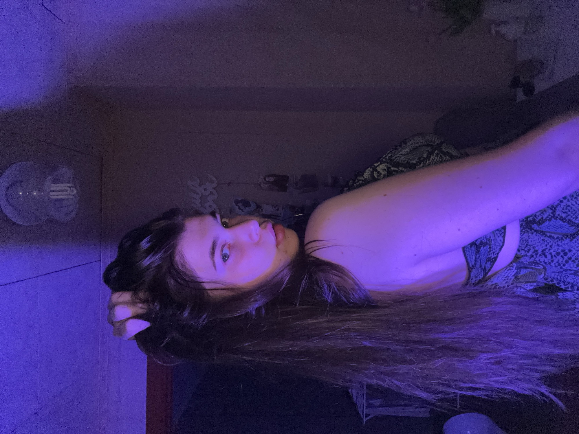 A woman with long brown hair is lying down, her head resting on her hand. She is wearing a patterned top. The environment is dimly lit with a blue light. There are household objects in the background, including what appears to be a light fixture on the wall. It is not possible to determine the location as Мещёры, Russia with the given information.