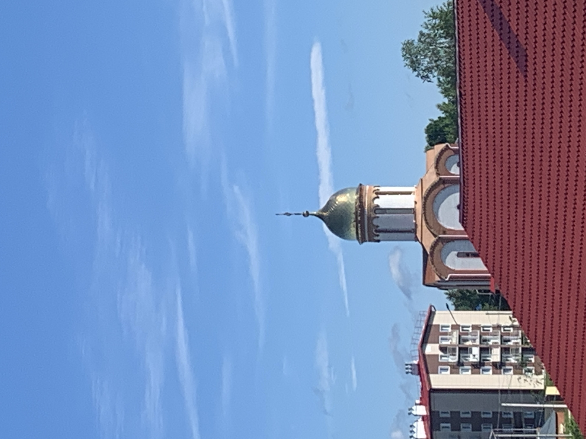 The image shows a view of the city of Volchansk, Russia, on a clear day with a blue sky and scattered clouds. In the foreground, a red-tiled roof dominates the lower portion of the frame. Behind the roof, a modern apartment building with a beige and brown facade, featuring multiple balconies and white-framed windows, stands on the left. To the right of the apartment building, a church with a distinctive golden dome and white and beige architectural details is visible. The church has arched windows and a tall, slender spire topped with a cross. Lush green trees are seen behind the church. The overall scene depicts a residential area in Volchansk with a mix of modern architecture and religious buildings under a bright sky.