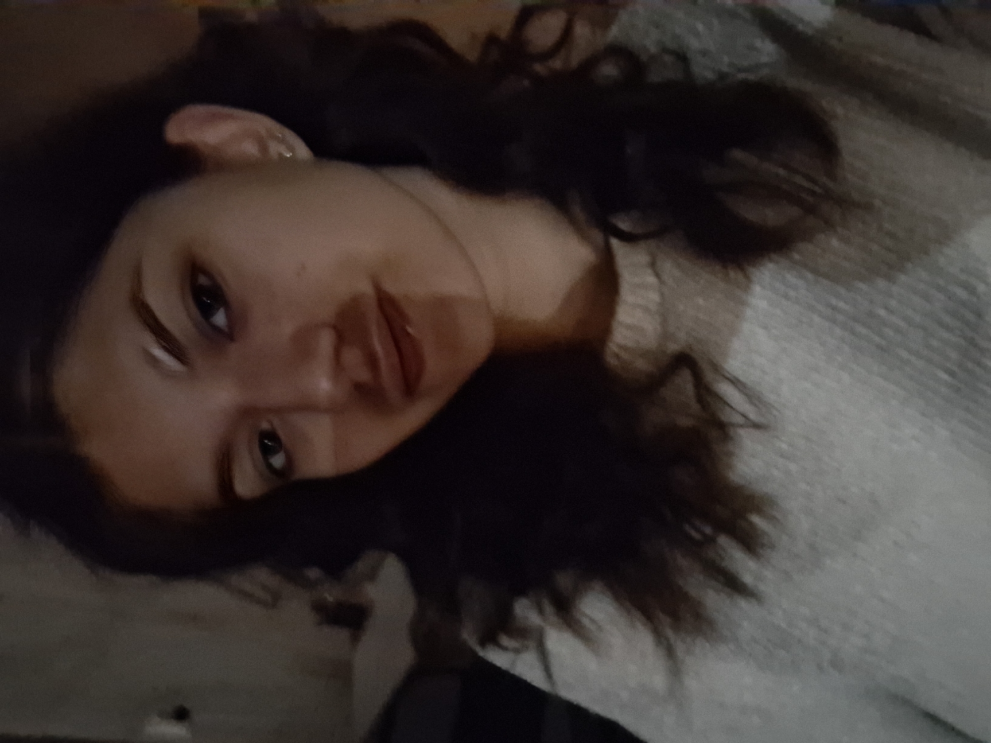 A woman is lying down, with her head slightly tilted towards the camera. She has dark hair, light skin, and is wearing a light-colored sweater. The background is blurry and dark. The location is unknown.