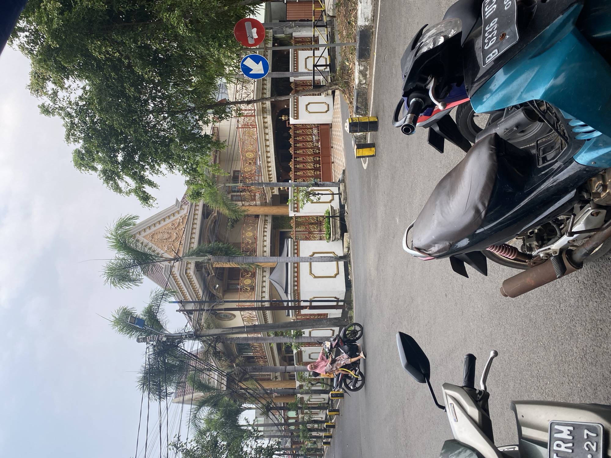 In the city of East Jakarta, Indonesia, a street scene unfolds with a large, ornate house as the backdrop. The house features a light-colored facade with elaborate gold trim on its balconies and roofline, accented by palm trees lining its front. A woman wearing a pink hijab and a patterned outfit rides a motorcycle with a child on the back down the paved street. Two motorcycles are parked in the foreground, one predominantly silver and the other a combination of black and teal. Traffic signs are visible on the right side of the road, indicating traffic direction and a prohibition. The sky is overcast, and the overall environment suggests a residential urban setting.