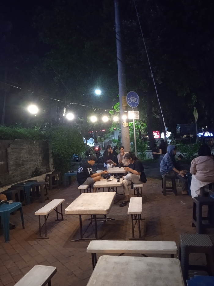 The scene is a nighttime outdoor setting with several people sitting around tables at a cafe or food stall. The location is on a street in Surakarta, Indonesia (latitude: -7.57, longitude: 110.82). The tables are made of metal and have white tabletops. Some of the chairs are metal folding chairs and some are wooden. The people are casually dressed in casual attire. The scene is lit by string lights and streetlights. There is a brick wall to the left of the scene and a green metal street sign for bikes to the right. Behind the tables are trees and greenery. There are people walking and driving in the distance. The people at the table are interacting with each other. The tables are set with drinks and food. The atmosphere is casual and relaxed. One person is using their phone. The scene is captured at night.