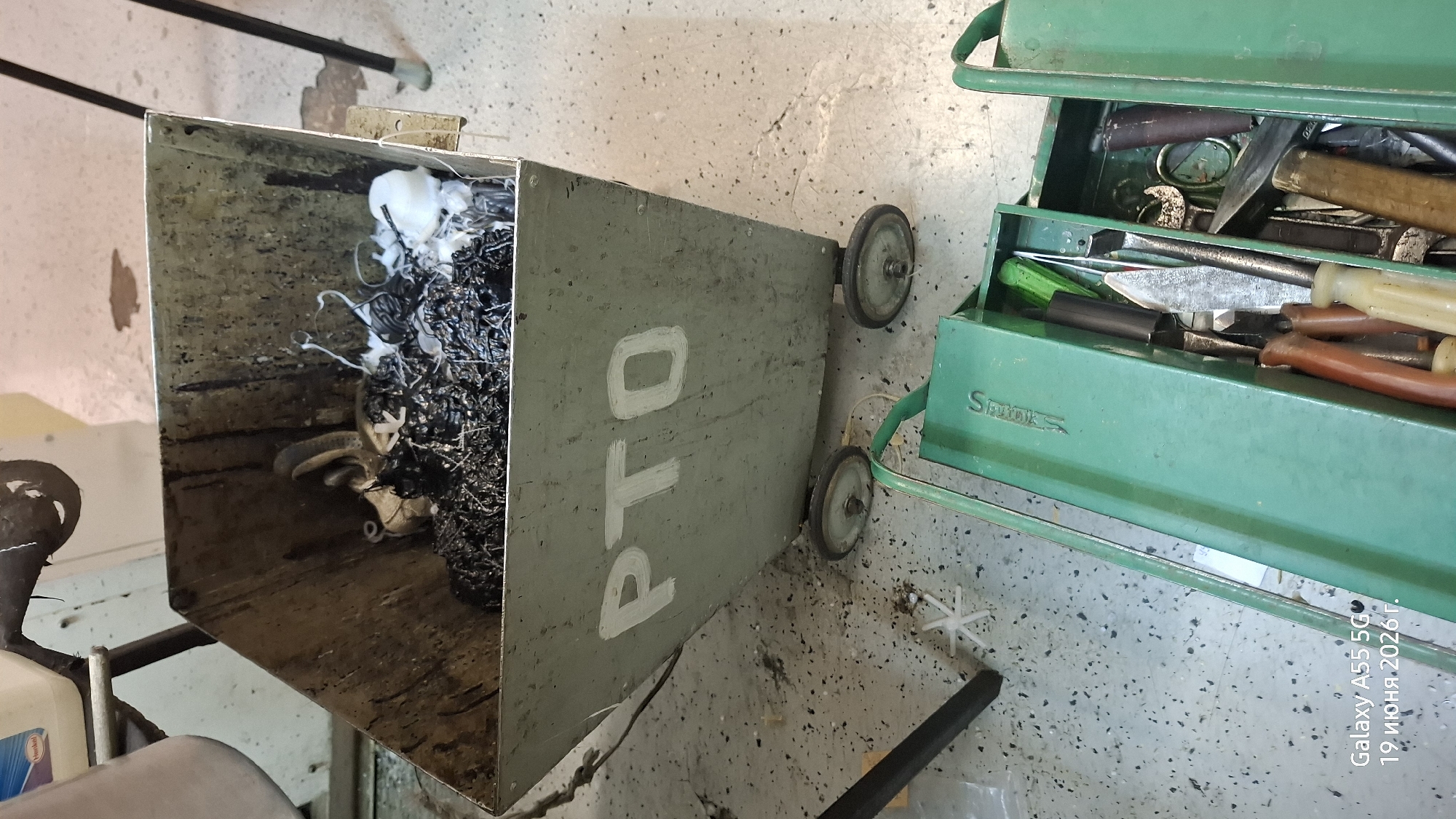 The image depicts a workshop scene, likely in Rostov-on-Don, Russia. The primary objects are a large, grey, metal bin filled with what appears to be metal shavings and some white material, and a green metal toolbox on wheels. The bin has "PTO" painted in white on its side. The toolbox contains various tools, including hammers, chisels, and screwdrivers. The floor is a speckled concrete surface, typical of an industrial or garage setting.
The lighting suggests it is daytime, and there are no indicators of specific weather conditions. The scene is static, with no discernible activity or event taking place beyond the potential storage of materials and tools. The overall impression is one of a functional, perhaps slightly neglected, workspace.
Visible text includes "PTO" on the bin, and the brand name "Snap-on" is subtly embossed on the toolbox. Additionally, a timestamp in Russian is visible in the bottom right corner: "Galaxy A55 5G, 19 июня 2026 г." (Galaxy A55 5G, June 19, 2026).