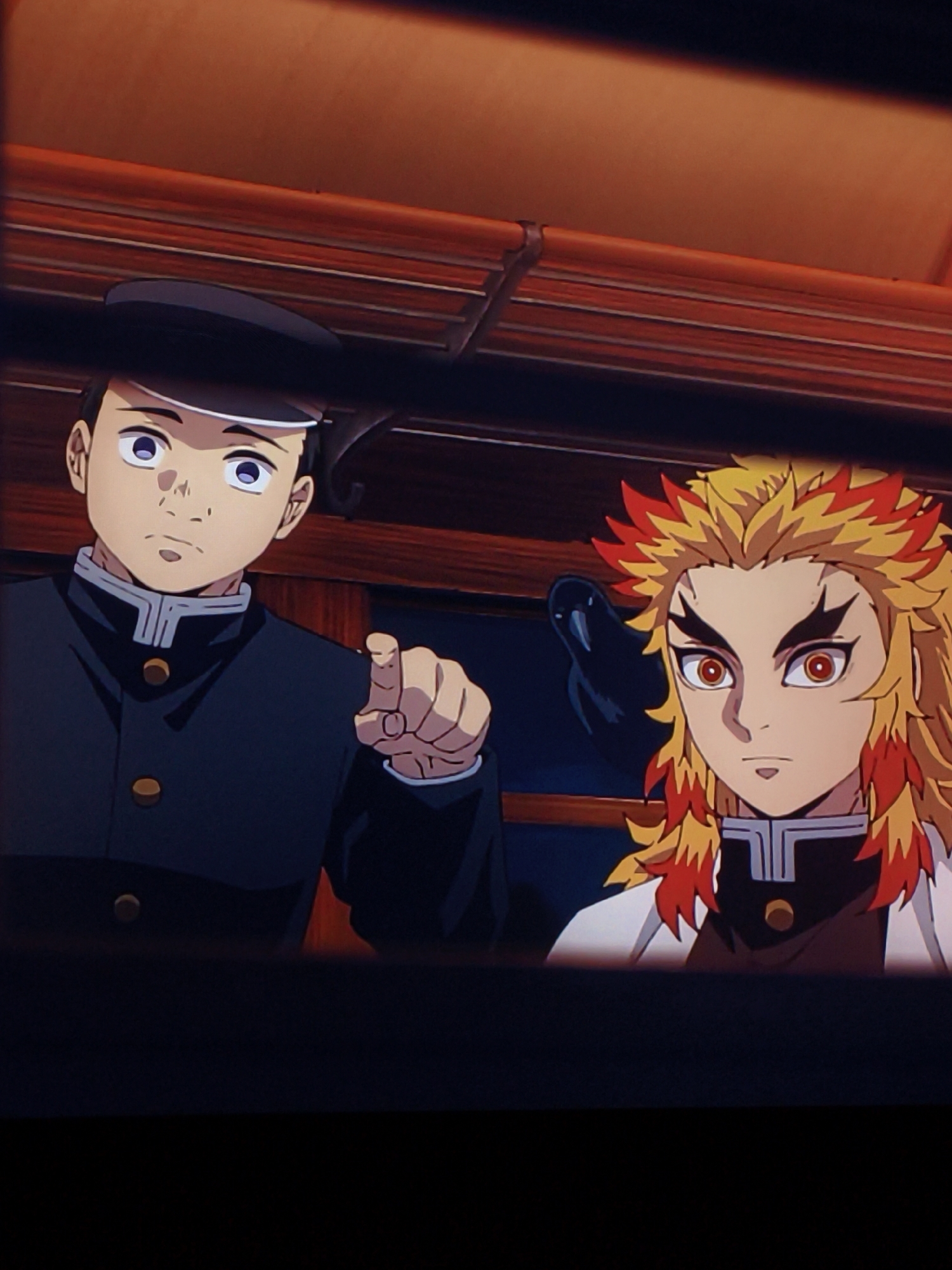 This is a close-up, vertically oriented, slightly blurry, animated image of two young male anime characters in what appears to be an indoor setting, possibly a train carriage. The character on the left wears a dark uniform with a peaked cap and points forward with his right index finger. He has short dark hair, large purple eyes, and a neutral expression. The character on the right has distinctive flame-like, spiky orange and yellow hair, large red eyes, and wears a dark uniform with white trim. He looks towards the viewer with a serious expression. The background consists of dark, wooden-paneled walls with horizontal slats, suggesting an interior with vintage or traditional decor. The overall lighting is dim. There is no discernible connection to the city of Vladivostok, Russia, or any specific urban environment.
