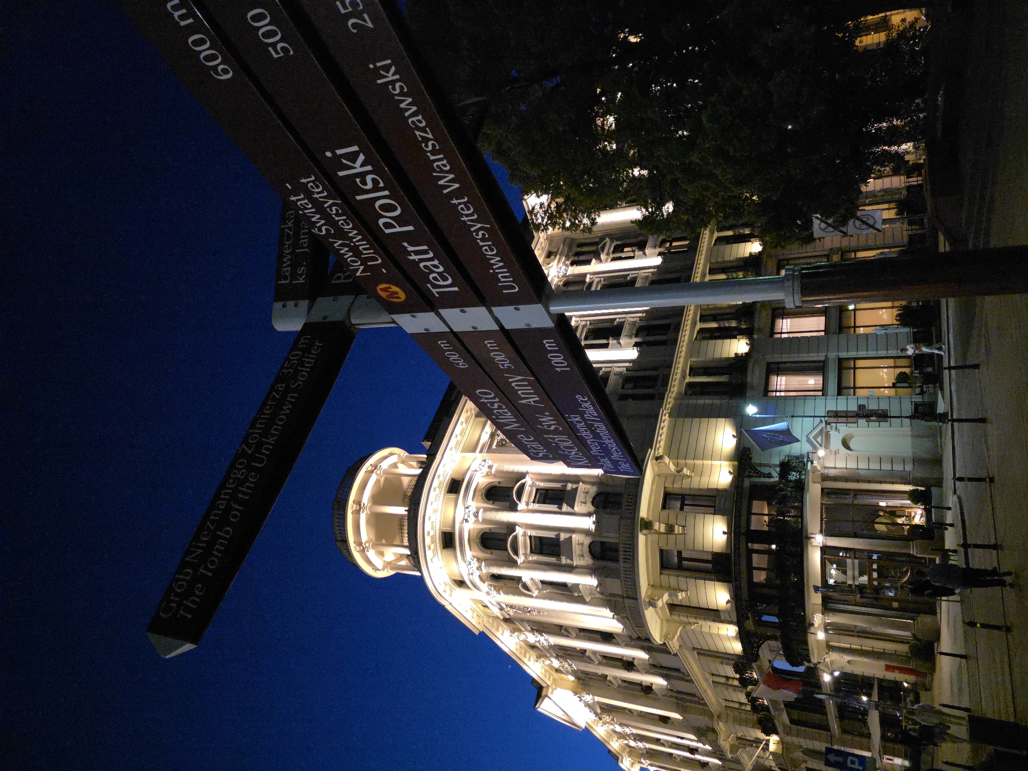 The image depicts a night scene in Warsaw, Poland. A signpost points to various landmarks: "Grób Nieznanego Żołnierza" (The Tomb of the Unknown Soldier, 350 m), "Nowy Świat - Uniwersytet" (New World Street - University, 600 m), "Teatr Polski" (Polish Theatre, 500 m), and "Uniwersytet Warszawski" (University of Warsaw, 25 m). Another sign points toward "Stare Miasto Kościół Św. Anny" (Old Town St. Anne's Church) and "Pałac Prezydencki" (The Presidential Palace, 100 m). The Presidential Palace is visible, illuminated with lights. A tree stands nearby and people walk on the sidewalk next to the street. The sky is a deep blue.