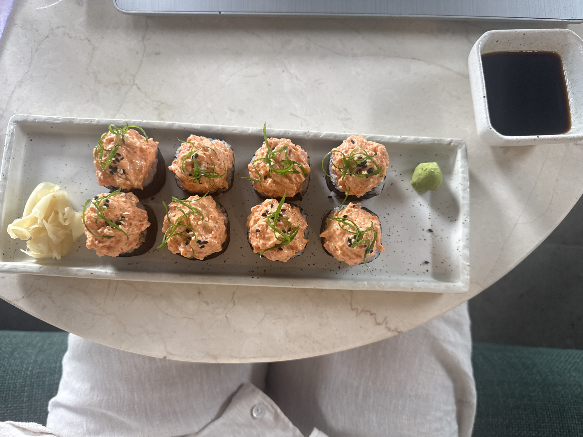 A top-down, eye-level shot captures a dish of sushi rolls served on a long, rectangular, speckled white plate. The plate contains eight sushi rolls, each topped with a dollop of orange-colored spicy tuna and garnished with thinly sliced green scallions and black and white sesame seeds. To the top left of the plate, a small pile of pickled ginger is visible. Below the sushi rolls, on the plate, a small mound of bright green wasabi sits. To the bottom right of the plate, a small square white dish holds a dark liquid, presumably soy sauce. The plate rests on a light-colored, possibly marble, table. To the left of the table, a portion of a person's torso is visible, clad in a light-colored linen shirt with a button detail. Further to the left and in the background, a sliver of a dark green chair can be seen. The overall lighting is soft and natural, suggesting an indoor setting. The image does not contain enough information to determine the specific location in the city of Ubud District, Indonesia.