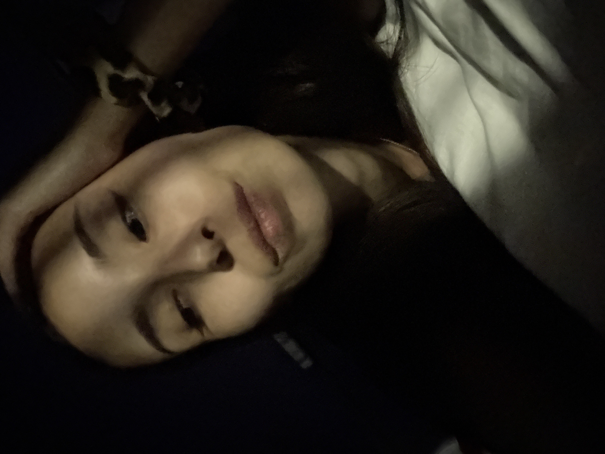 A dark, close-up, indoor selfie is taken by a young woman with dark hair and brown eyes. She is wearing a white t-shirt and has her right hand on her forehead. A leopard print scrunchie is visible on her left wrist. The background is dark, with a hint of a dark blue or black surface on the left side of the image. The lighting is dim, casting shadows across her face and the fabric of her shirt. The image appears to have been taken at night. There are no discernible architectural features or environmental clues to indicate the location is specifically Doma 1096 Km, Russia.