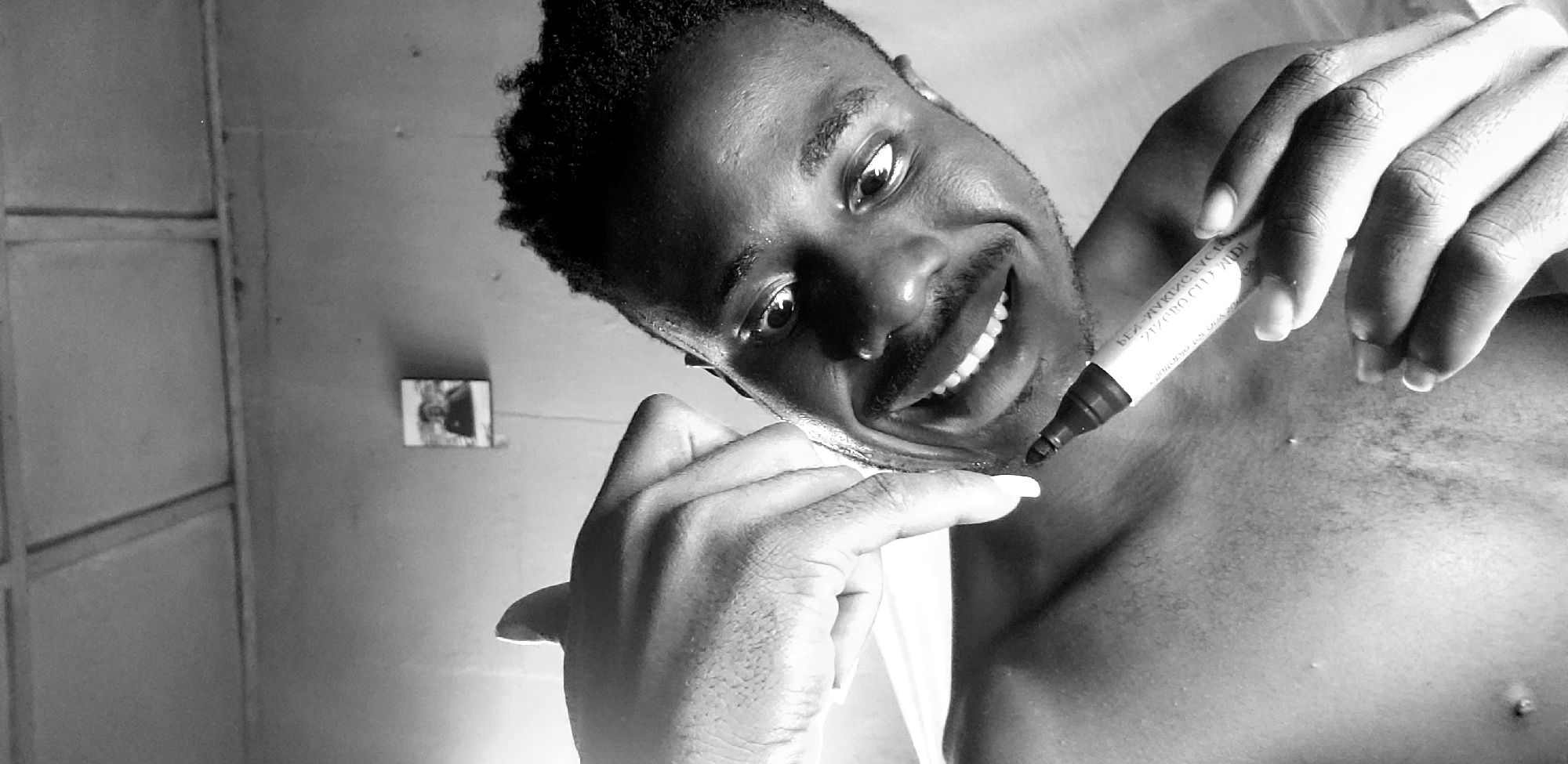 A black and white, close-up, eye-level, selfie-style shot shows a young Black man smiling widely while holding a marker to his chin. He is shirtless and looking directly at the camera. His right hand is raised, with his thumb and index finger extended and pointing towards his face, appearing to be in the process of applying something to his chin with the marker. The background is a textured wall with a ceiling visible at the top of the frame. A small framed picture is mounted on the wall behind him. The image appears to have been taken indoors, possibly in a residential setting. No specific location in Narayi, Nigeria is identifiable.
