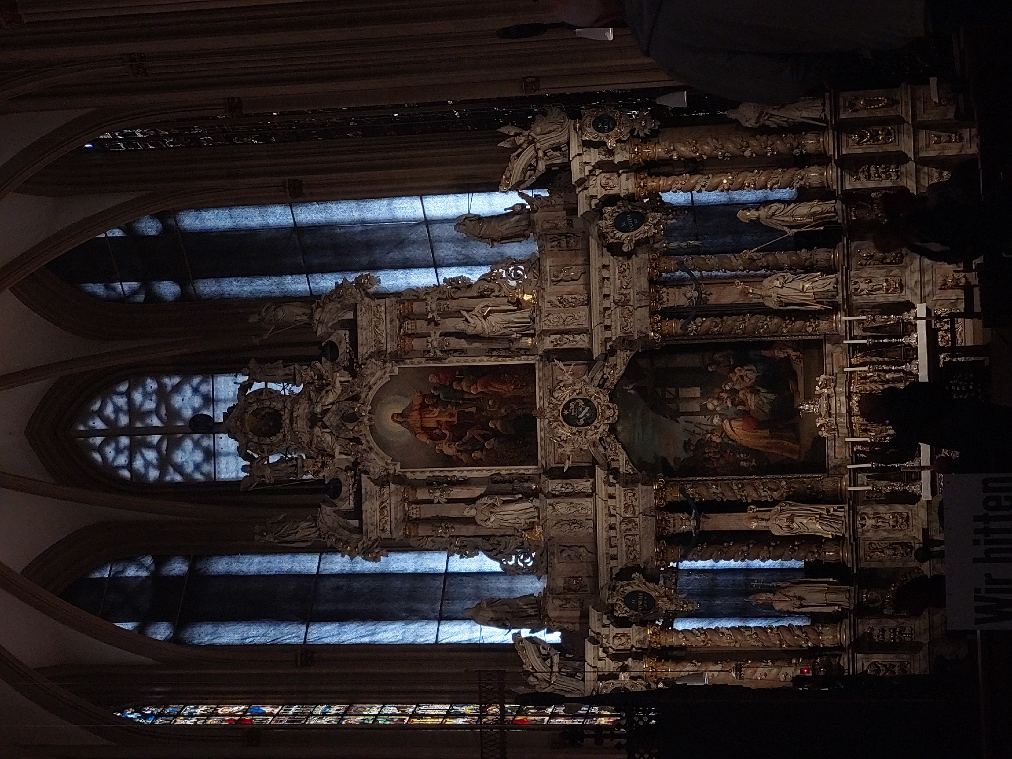 The image depicts the interior of a grand church, likely Saint Mary's Cathedral in Erfurt, Germany, given the prominent altar and architectural style. The central focus is an ornate, multi-tiered altar, rich with intricate carvings and gilded details. Above the altar, a large painting depicts a religious scene, possibly Jesus Christ with followers, illuminated by soft light. Flanking the painting are statues of religious figures integrated into the altar's structure. Several large stained-glass windows with arched tops are visible in the background, casting a cool, blue light into the dimly lit interior. The architecture exhibits Gothic elements, with ribbed vaults visible in the ceiling. In the foreground, a sign with German text, "Wir bitten," is partially visible, suggesting a place where donations are requested. Silhouetted figures of people are present, facing the altar, indicating the space is in use as a place of worship or tourism. One figure in the lower right corner appears to be an adult looking towards the altar. The overall atmosphere is solemn and reverent, characterized by the contrast between the darkness of the interior and the illuminated altar and windows.