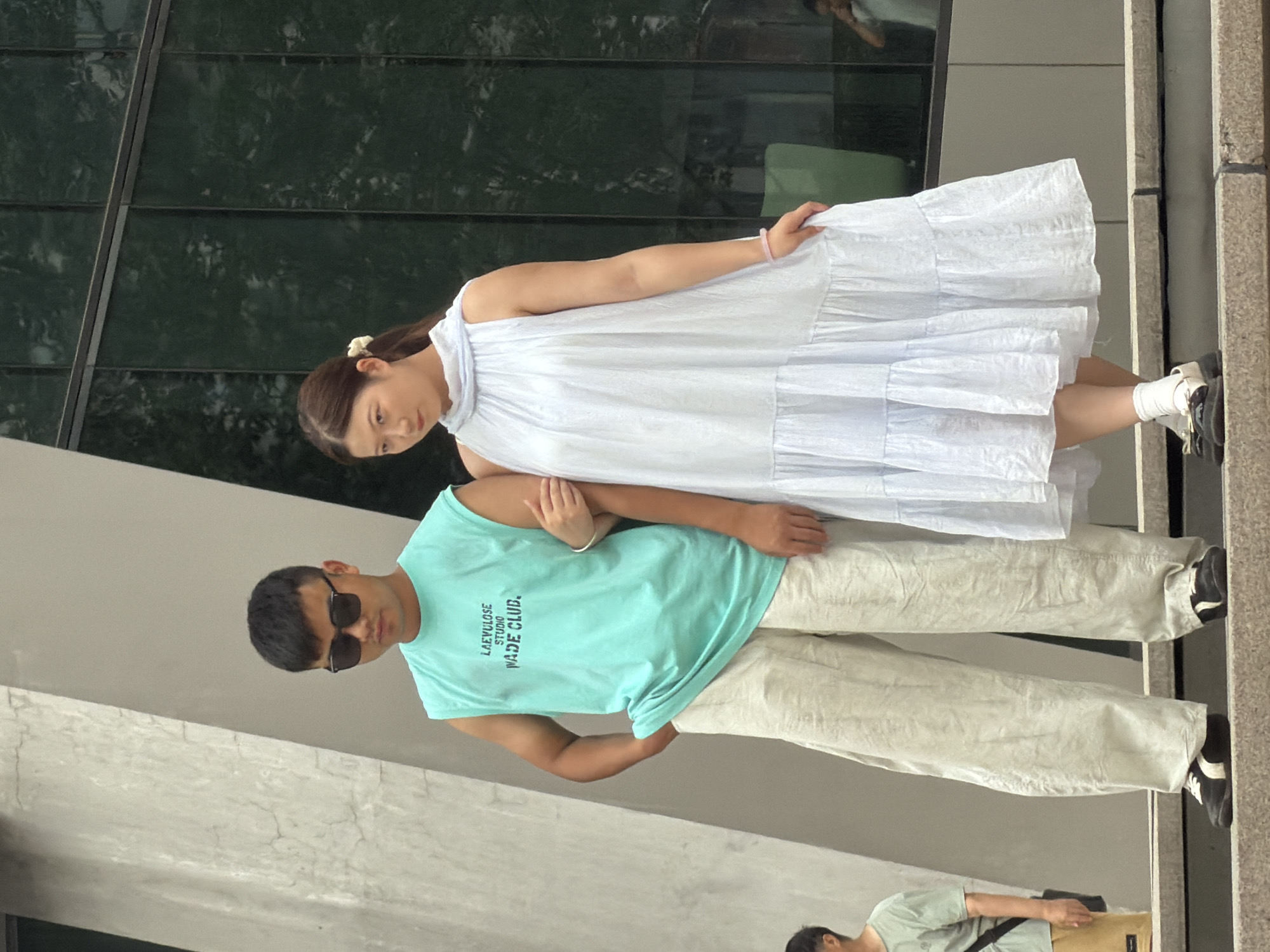 A man and a woman stand side-by-side on an outdoor staircase in Yuzhong District, China. The man, on the left, wears a sleeveless turquoise t-shirt with text and black sunglasses. He has on loose, light-colored pants and dark shoes with white stripes. His left hand is in his pocket, and his right arm is around the woman's shoulders. The woman, on the right, wears a pale blue, tiered dress with a high neckline. She has a pink bracelet on her left wrist and wears white socks with black and white loafers. Her left hand rests on her dress. Behind them is a modern building with dark, reflective windows, and a large, angled concrete structure. In the background, a person in a green shirt is partially visible.