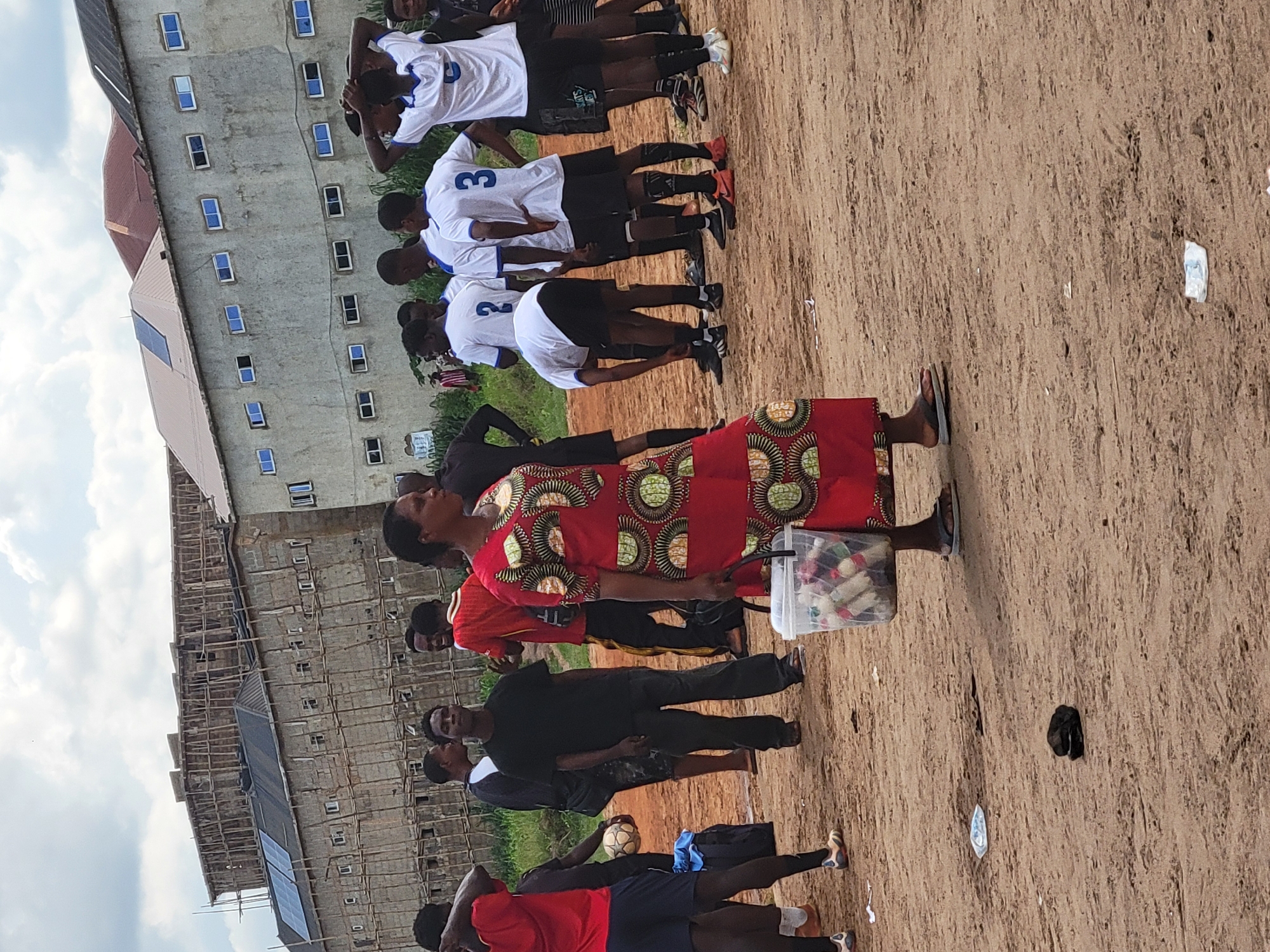 In Egbagu, Nigeria, a group of young men in white and blue soccer uniforms stand around on a dirt field. A woman in a vibrant red and green patterned dress walks past them, carrying a clear plastic bucket filled with small items. Behind the group, a multi-story building under construction with scaffolding and solar panels on its roof dominates the background. The sky is overcast with scattered clouds.