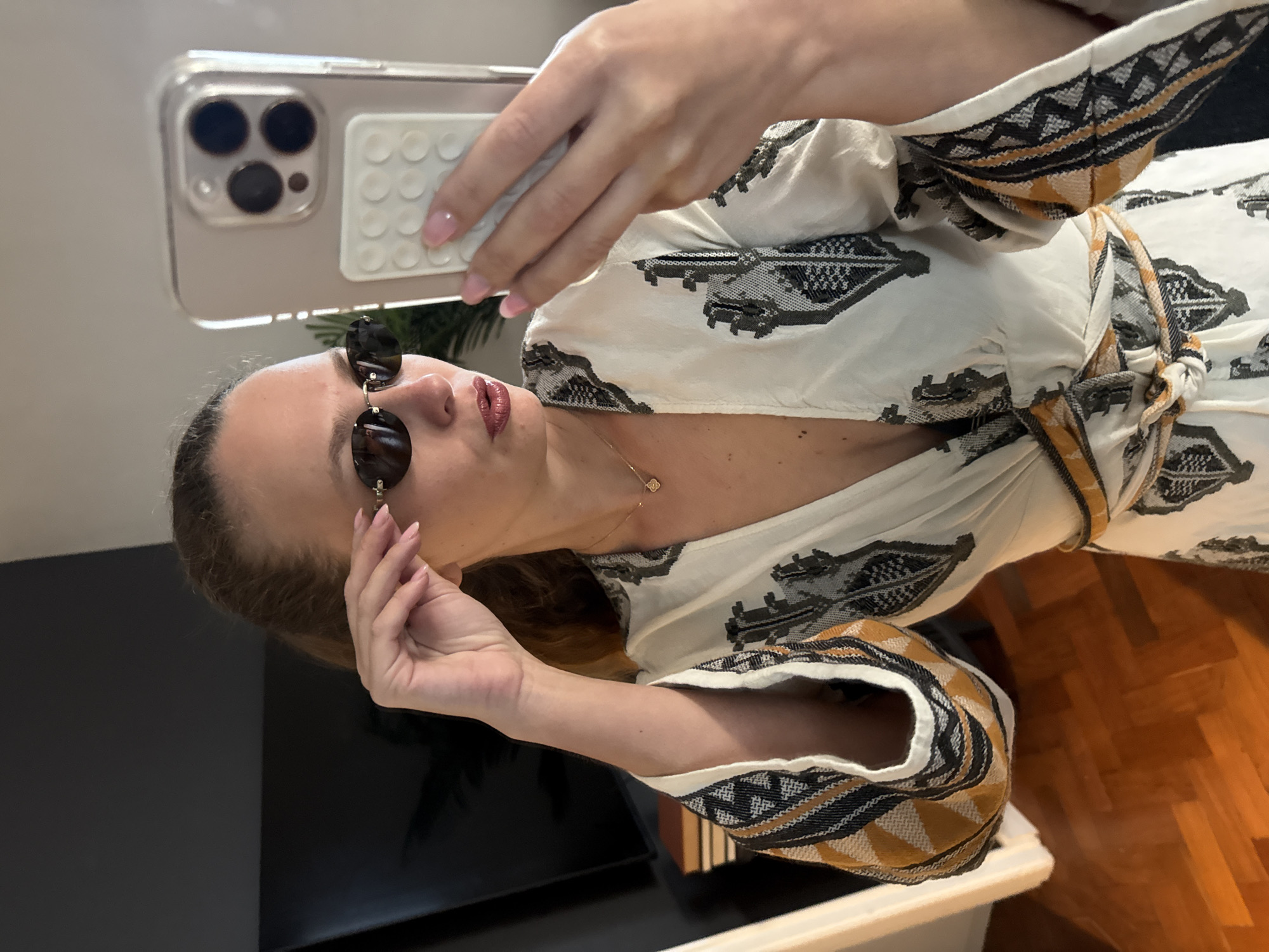 A woman is taking a selfie with a smartphone. She is wearing a white dress with a tribal pattern and black sunglasses. The background includes a black surface, a wooden floor, and a glimpse of a houseplant. There is no information to connect the image to Cape Town.