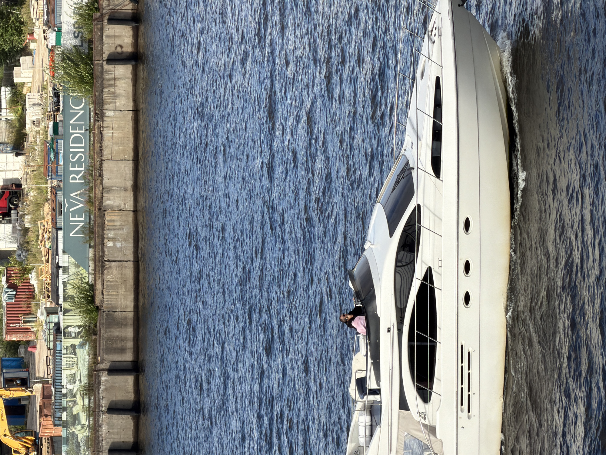 A white motor yacht is cruising on a body of water, possibly a river or canal. The yacht has a sleek design with large windows and a flybridge. On the flybridge, two individuals are visible: one person, possibly a woman, is wearing a pink top and dark trousers, while the other person, possibly a man, is wearing a black top and sunglasses, appearing to be steering the boat. In the background, a concrete embankment lines the shore. Above the embankment, a mural is painted on shipping containers, depicting figures in a domestic setting. Further back, industrial elements are visible, including a yellow excavator, red shipping containers, and scaffolding. A sign on the containers reads "NEVA RESIDENCE". The water is choppy, suggesting movement. The overall scene appears to be an urban waterfront area in Saint Petersburg, Russia, likely along the Neva River or one of its tributaries, given the "Neva Residence" sign.