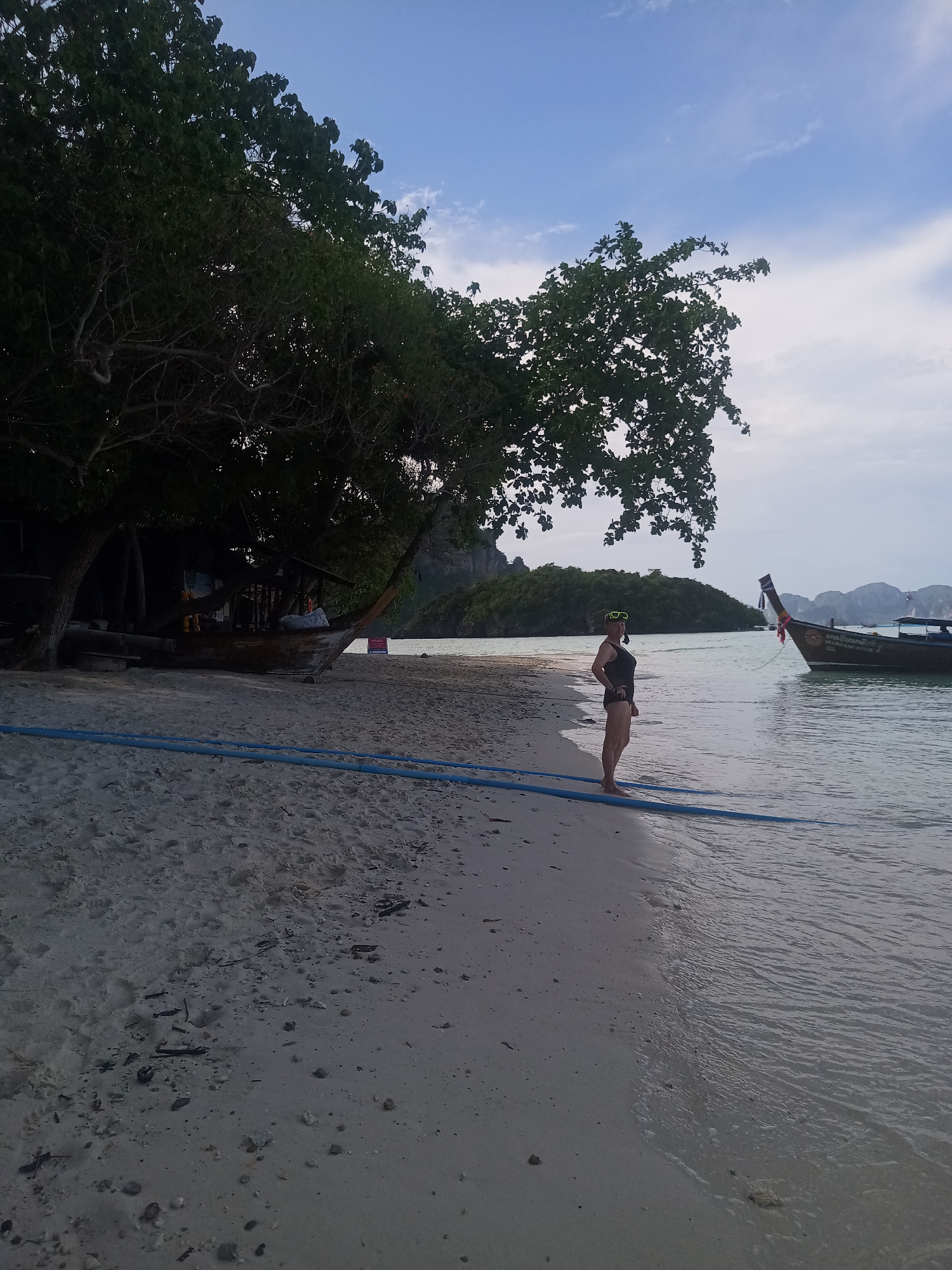The image shows a beach scene in Ao Nang, Thailand. A woman in a black swimsuit and goggles stands at the water's edge. A longtail boat is anchored in the shallow water near her. The beach is sandy with some debris. In the background, there is a tree that provides shade and a boat resting under it. There is an island in the distance, and the sky is a mix of blue and overcast. A blue pipe lies on the sand.