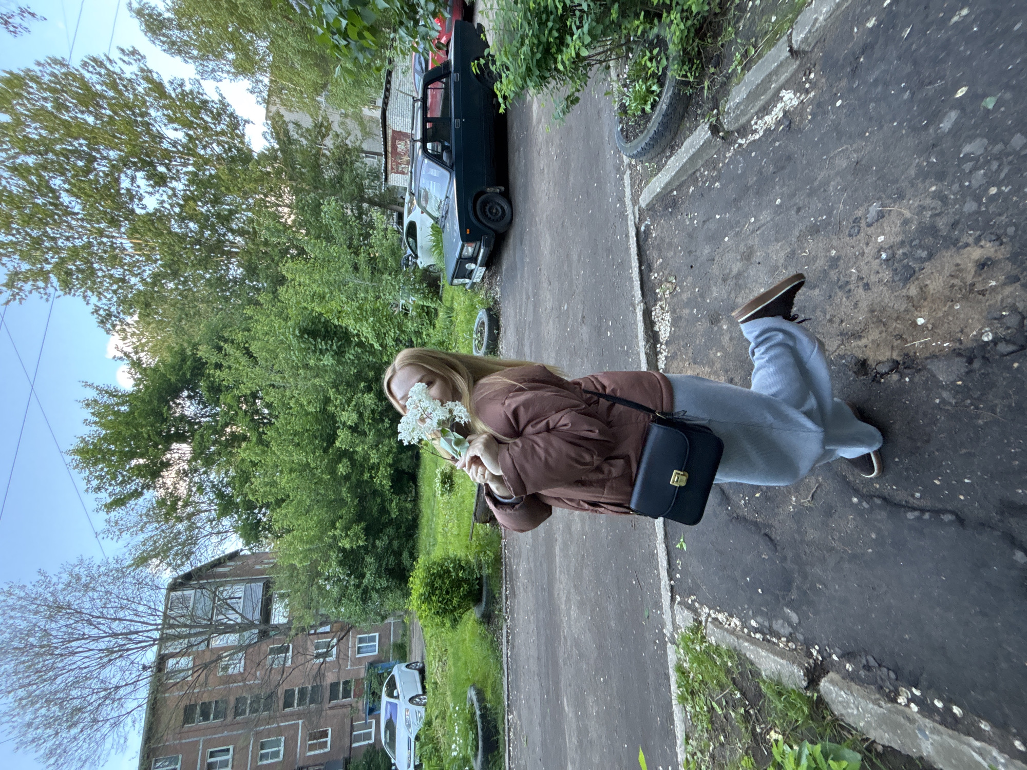 A young woman stands on a worn asphalt sidewalk in Rusanovo, Russia, holding a bouquet of white flowers to her face. She wears a brown puffer jacket, light blue pants, and dark-colored shoes. A black purse hangs from her shoulder. The background includes a residential building, green trees, and several parked cars, including a dark green Lada. The sidewalk is bordered by a concrete curb and scattered with fallen flower petals. A tire rests near a patch of grass and foliage.