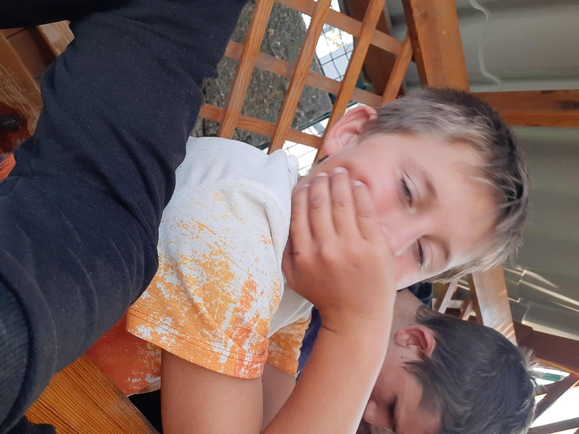 In this close-up, slightly tilted photograph, two individuals are partially visible. The foreground is dominated by the arm and torso of one person wearing a black long-sleeved garment. Next to this, the torso and arm of another individual are visible, wearing a white t-shirt with an orange and yellow abstract pattern. Their right hand is covering their mouth and nose area. The focus shifts to the face of a third person, whose head is turned to the side, with their eyes closed and right hand partially obscuring their face. They appear to be very young.
The setting seems to be outdoors, possibly under a wooden structure with latticed beams. Sunlight filters through, suggesting it is daytime. The lighting is bright, creating some harsh shadows. There is no visible text in the image. The overall impression is one of a candid, informal moment, perhaps of children playing or interacting.