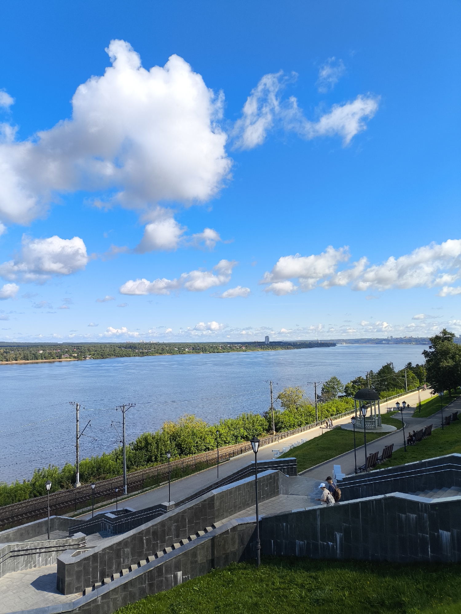 This is a wide-angle, eye-level outdoor shot of a riverfront park area in Perm, Russia, on a clear, bright day with a vibrant blue sky dotted with fluffy white clouds. In the foreground, a wide, tiered stone staircase with dark gray cladding descends from the right into the lower portion of the frame. Two individuals, a male and a female, are walking up the staircase, the male wearing a backpack. The staircase is bordered by manicured green grass on its right side and stone-paved walkways on its left.
Beyond the staircase, a paved park pathway curves along the riverbank, lined with benches and lampposts. A decorative gazebo with a green roof sits on the left side of the pathway. Further to the left, a set of train tracks runs parallel to the river, bordered by dense green foliage and trees.
The wide, blue river dominates the midground, reflecting the bright sky. On the far bank of the river, a dense line of green trees and vegetation stretches across the horizon, with scattered buildings and a distant tall structure visible. The overall impression is one of a well-maintained public space on a beautiful summer day.