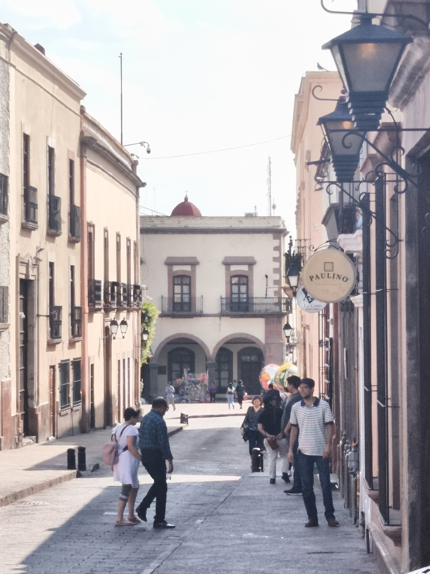 This street scene is in the city of Querétaro, Mexico, with cobblestone streets lined by pale pink buildings. Three old-fashioned street lamps are on the right, affixed to the buildings with decorative ironwork. One sign reads "Paulino." In the foreground, a woman in a white dress and pink backpack walks with a man in a blue plaid shirt. Further down the street, a group of people is walking, including a man in a striped shirt. In the distance, an arched building with a red dome is visible. People are milling about in that area.