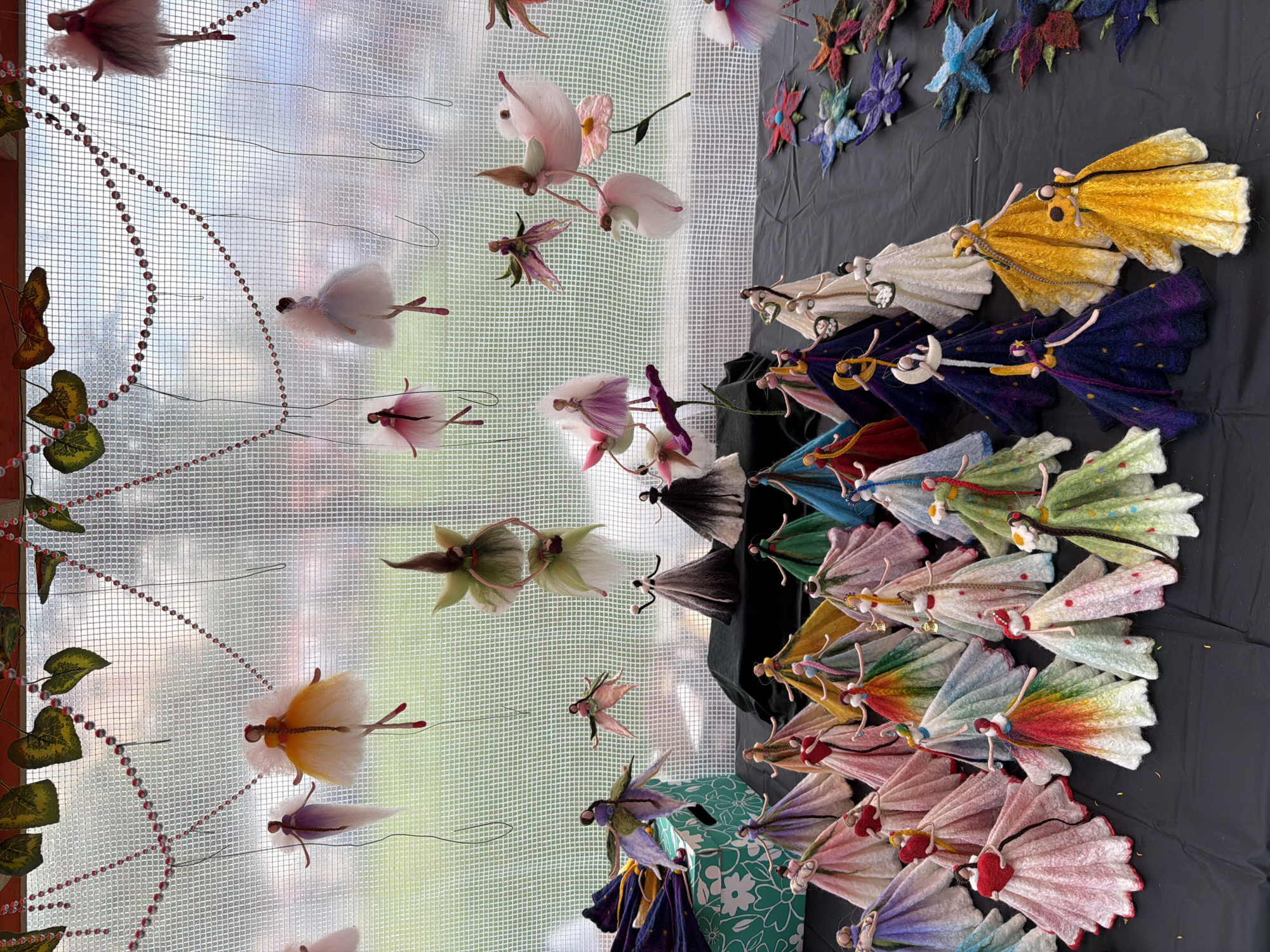 The image depicts a display of numerous handmade fairy figurines, crafted from fabric and other materials, arranged on a black surface. The figurines vary in color and design, many featuring voluminous, layered skirts and delicate, articulated limbs. Some fairies are suspended from a netting background by thin threads, while others stand upright on the surface. The background consists of a light-colored netting with a blurred outdoor scene visible through it, possibly suggesting a market stall or outdoor exhibition. Decorative beads and faux leaves are strung across the top of the netting. The overall impression is a whimsical collection of artistic creations, possibly for sale or exhibition. The location is identified as Vracar Urban Municipality, Serbia.