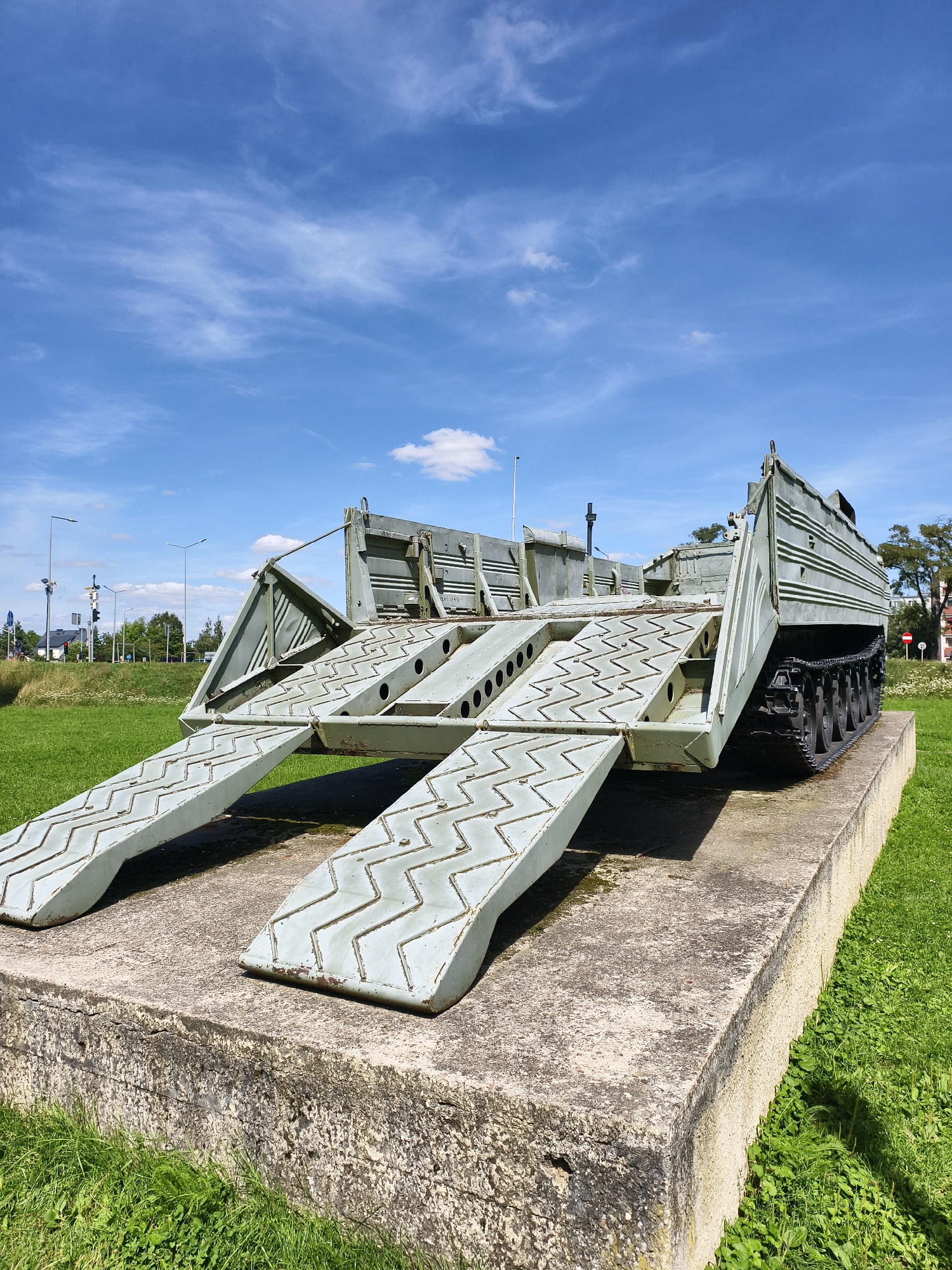 An outdoor, low-angle, medium shot captures a static, olive-drab military bridge-laying vehicle positioned on a concrete pedestal in Tczew, Poland. The vehicle, which appears to be a recovery or engineering vehicle, is a segmented bridge transporter with two ramps extending forward at an upward angle. The ramps have a distinct zigzag texture for traction and are supported by metal beams. The main body of the vehicle is broad and boxy, with visible tracks and some external machinery. Behind the vehicle, a street with traffic lights and power poles is visible, along with trees and a grassy field. The sky above is a bright blue with scattered white clouds. The scene is set on a sunny day.