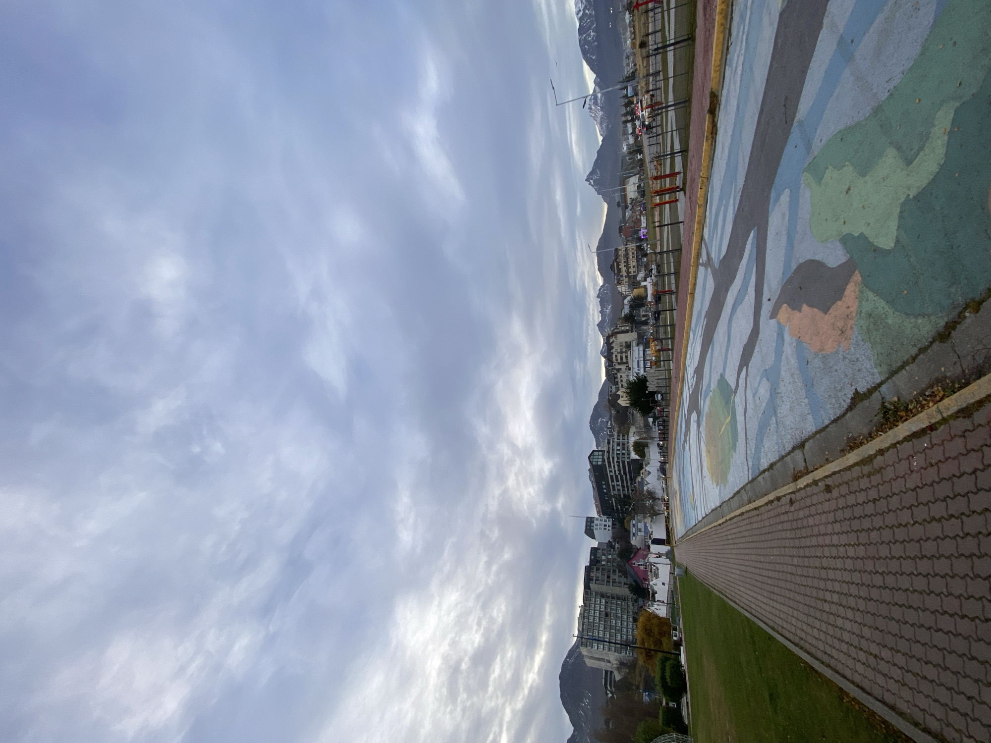 The image depicts a cityscape of Ushuaia, Argentina, seen under a cloudy sky. The cityscape is oriented horizontally, with buildings of varying heights clustered along the edge of mountains. A paved walkway, with a brick pattern, runs along the right side of the frame, adjacent to a grassy area and a section painted with abstract shapes. In the background, snow-capped mountains are visible. The image's perspective suggests it was taken from an elevated position, possibly along a waterfront or a park area that offers views of the city and the surrounding landscape.