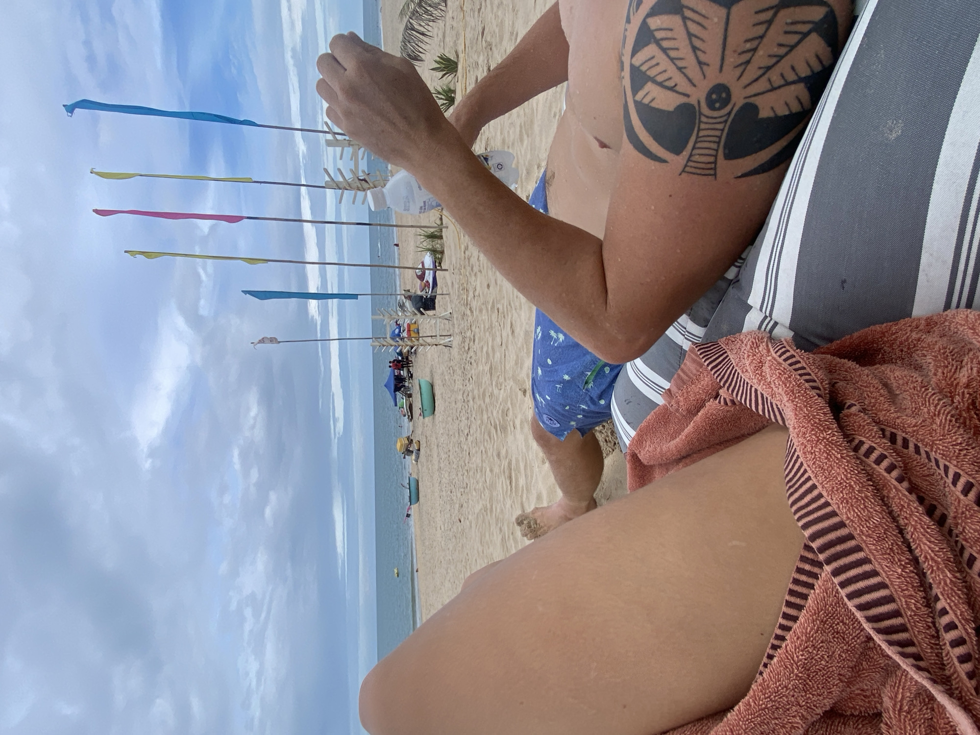 The image shows a partial view of a person at a beach in what appears to be Phu Thuy Ward, Vietnam. The beach has several colorful flags blowing in the wind, and the sea and a cloudy sky are visible in the background. In the foreground, there is a person sitting in a beach chair with a towel covering their lap, a man is sitting next to the person in blue swim trunks. The man has a tattoo on his upper arm.