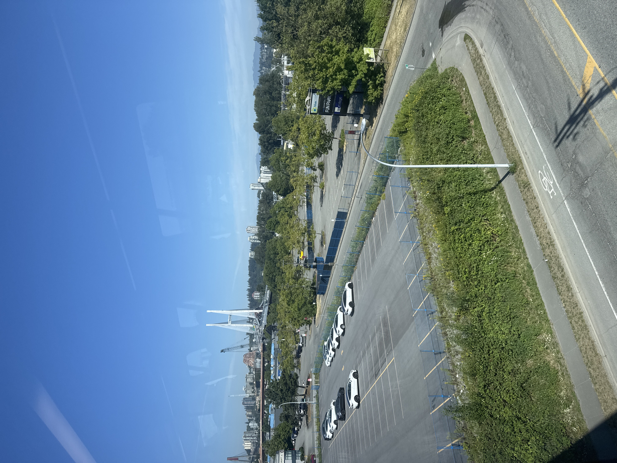 The image shows a high-angle view of a parking lot and surrounding area in Surrey, Canada, under a clear blue sky. In the foreground, a paved parking lot is marked with white lines, and several white cars are parked in rows. Beyond the parking lot, there is a road with a bicycle lane painted on it, bordered by a curb and a grassy area with dense green bushes. In the middle ground, a large empty parking lot is visible, surrounded by trees and a blue fence. Further in the distance, a cityscape is visible with buildings and a large cable-stayed bridge. The overall environment appears to be an urban or suburban setting with a mix of commercial and residential structures, interspersed with green spaces and infrastructure. The lighting suggests it is daytime, likely late morning or early afternoon, given the bright sunlight and shadows.
