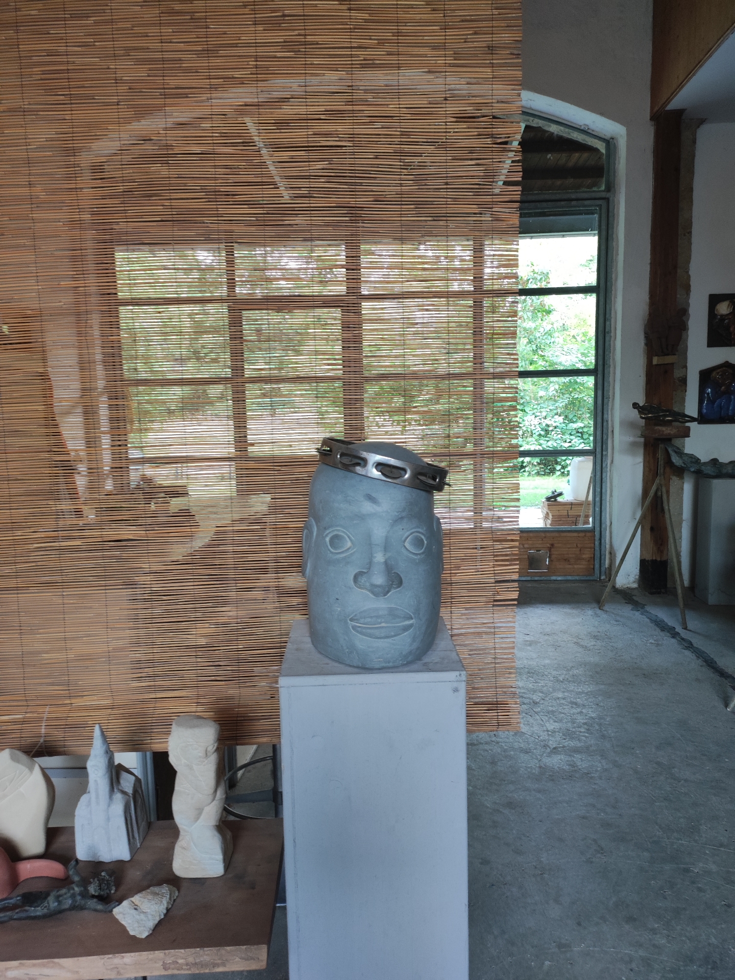 A grey stone head sculpture, adorned with a metal tambourine on its head, stands on a white pedestal in what appears to be an artist's studio in Dresden, Germany. The sculpture is positioned in front of a bamboo blind covering a window. Through the window, greenery and an outdoor scene are visible. To the left of the stone head, on a wooden surface, are several smaller sculptures, including a white, architectural-style piece and another abstract stone carving. The floor is concrete. To the right of the main sculpture, a doorway opens to an outdoor area with some wooden crates and vegetation. A wooden easel-like structure with some art pieces on it is also visible in the background. The overall atmosphere suggests a creative workspace.