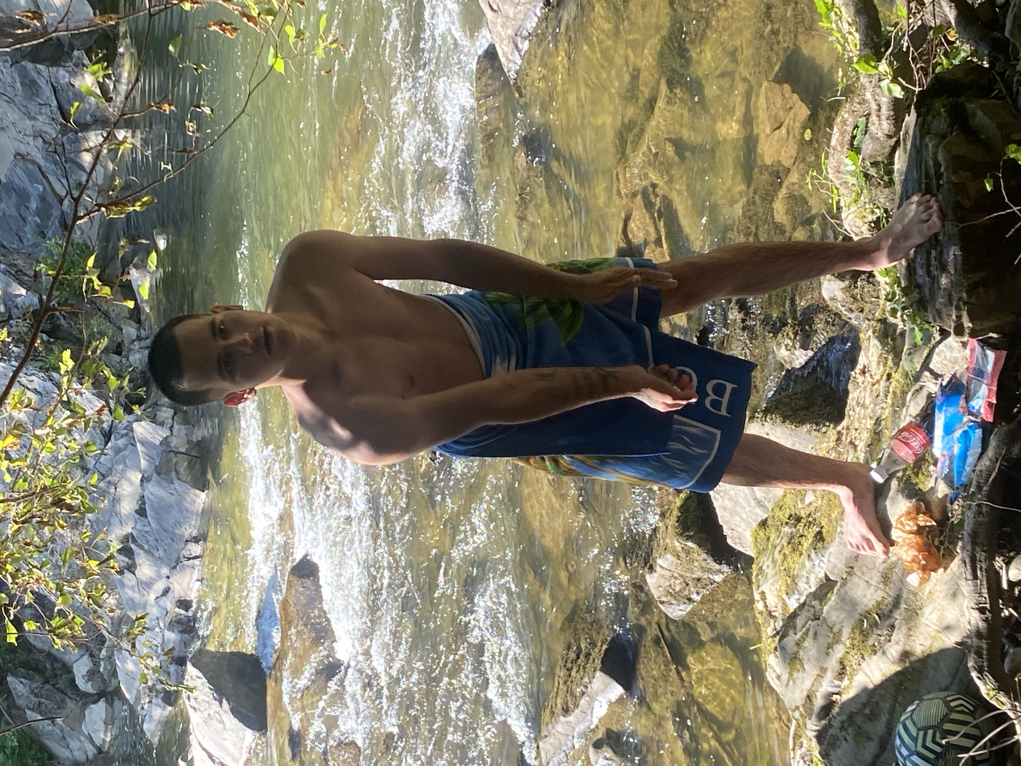 A shirtless young man stands on rocks beside a shallow, fast-flowing river with clear, green water. He is wearing a blue towel wrapped around his lower body. The riverbed is visible with submerged rocks. The surrounding environment consists of large, grey rocks, some covered with moss and vegetation. Overhanging branches with green leaves cast dappled sunlight onto the scene. In the foreground, on a large rock, are several items: a brown crumpled object, a red Coca-Cola bottle, and a stack of brightly colored snack bags. A soccer ball with a black and green pattern is partially visible to the left. The overall setting suggests an outdoor recreational area, possibly a riverbank in or near the city of Gumoshtnik, Bulgaria, with evidence of human presence and discarded items.