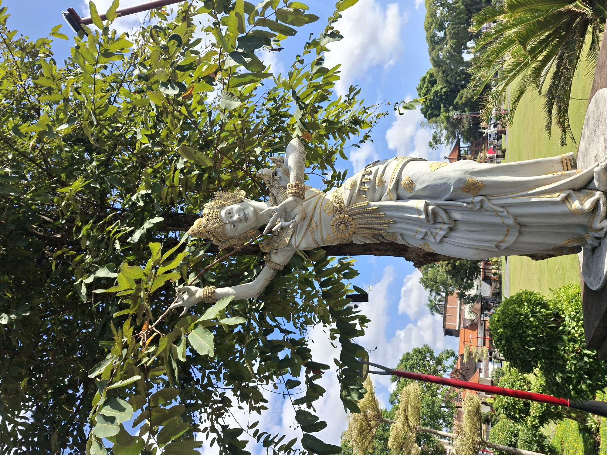 A full-shot, eye-level, outdoor, medium-angle, natural light photograph captures a stone statue of a Balinese dancer situated in front of a large tree with green foliage and a blue sky with scattered clouds. The statue, primarily white with gold accents, depicts a woman in traditional Balinese attire, including an elaborate headdress, ornate jewelry, and flowing garments. Her arms are raised, and her hands are positioned in a dance-like gesture. The statue is positioned on a stone pedestal. Behind the statue, a lush green lawn stretches towards a cluster of buildings with orange tiled roofs. Several trees, including palm trees, are visible in the background, along with a red pole with a black fixture. The overall scene suggests an outdoor public space, possibly a park or a cultural site, in Gianyar, Indonesia.