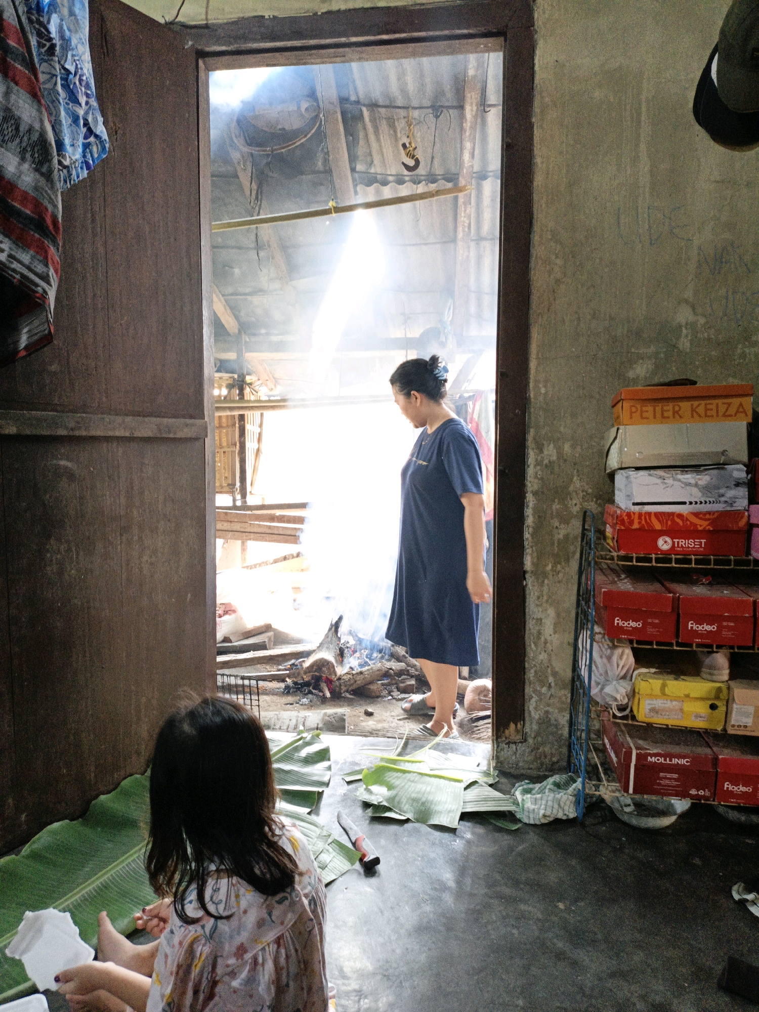 In a doorway in Bunut, Indonesia, a young girl sits on a mat of banana leaves, her back to the camera. She wears a patterned dress and holds a white object in her hands. To her right, a knife lies on the leaves. Beyond her, a woman in a blue dress stands in the entryway, looking into an outdoor space where smoke rises from a fire pit with burning wood. The scene is enclosed by rough walls and a wooden door frame. Shelves filled with shoe boxes are visible on the right.