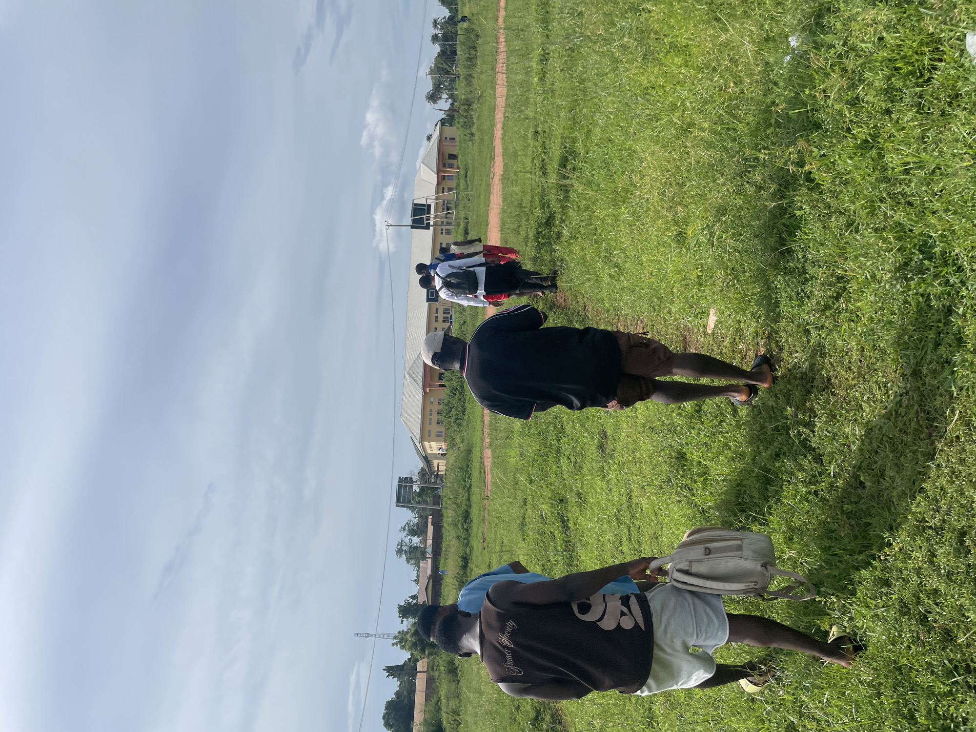 The image captures a group of people walking in a line across a sunlit green grass field in Ogbogbo, Nigeria. The perspective is from behind the group, with individuals moving away from the viewer along a dirt path that traverses the field.
In the foreground, a person is visible from the back, wearing a brown t-shirt with the text "Summer Society" printed on it, light-colored shorts, and carrying a beige backpack. Ahead of them, another individual wears a black t-shirt, dark shorts, and a white baseball cap. Further down the line, several more people are seen, some wearing white shirts, one in blue, and another in red.
The setting features a wide expanse of green grass. In the mid-ground, a metal-framed sign or board stands near the path. The background reveals light-colored buildings, possibly institutional or residential, with distinct roofs, under a clear, bright blue sky with scattered white clouds. Sparse trees and utility lines are also visible in the distance.
The scene is well-lit by natural daylight, suggesting it is mid-morning or early afternoon, with clear shadows cast by the people and structures. The overall impression is of a group undertaking a walk or procession through an open, possibly campus or community, area.