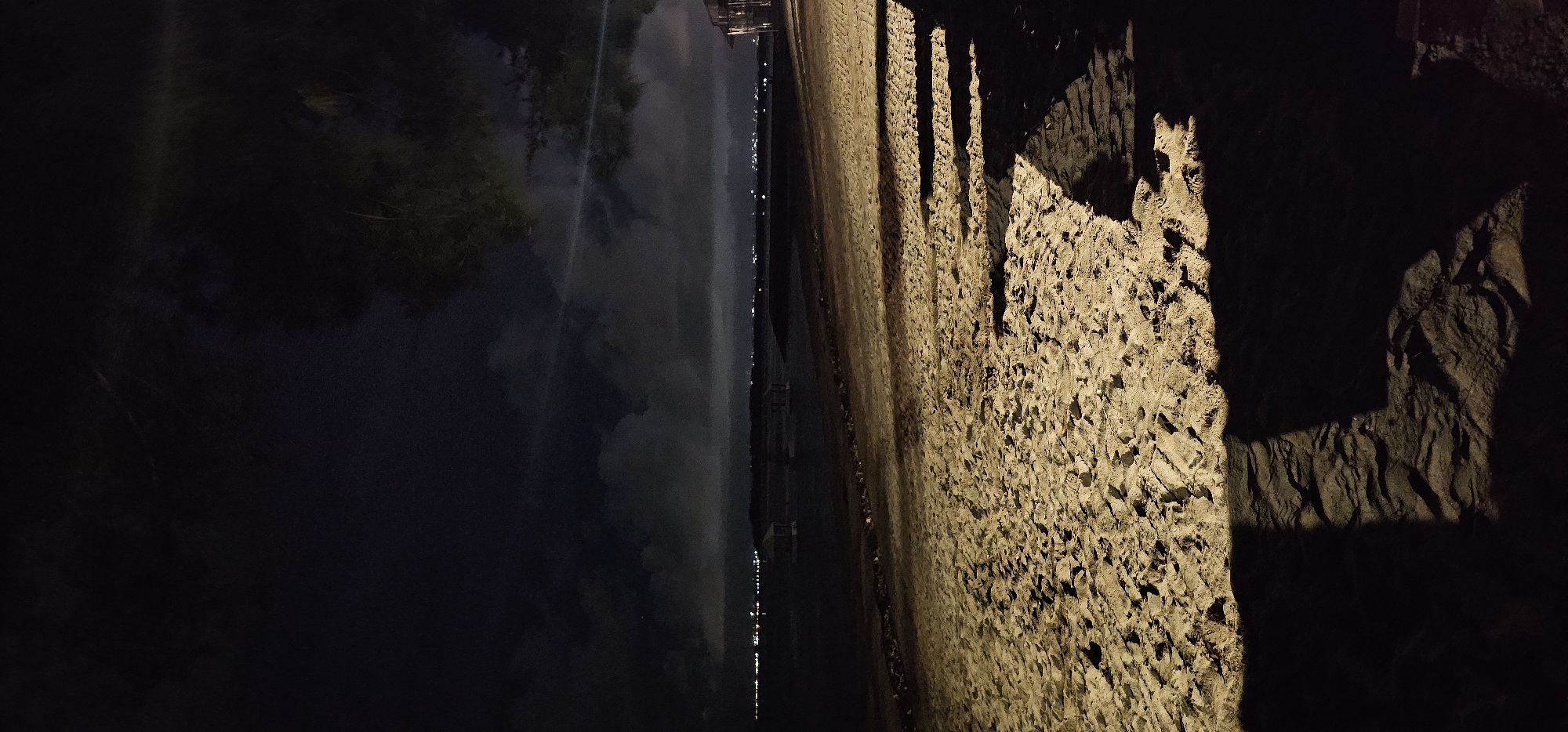 The image captures a nighttime scene along a body of water, likely a river or canal, in Denpasar, Indonesia. The sky is dark, indicating a clear night.
On the left side of the frame, a dark body of water reflects numerous small, bright lights stretching horizontally across the distant bank, suggesting a city or developed area. Above the water on the far left, dark, indistinct foliage or trees are visible, with their forms partially mirrored in the water's surface.
The right side of the image is dominated by a steeply sloped, rough-textured embankment or wall, possibly made of rock or coarse concrete. This embankment is brightly illuminated by an unseen artificial light source from the right, which casts long, prominent shadows across its uneven surface. These shadows reveal the contours and depth of the material. Several distinct, elongated vertical shadows are visible, implying objects or structures casting them from outside the frame.
No specific activities or people are discernible. The water appears calm, given the clear reflections. There is no visible text in the image. The overall atmosphere is tranquil and highlights the interplay of artificial light and shadow on the natural and man-made elements of the night landscape.