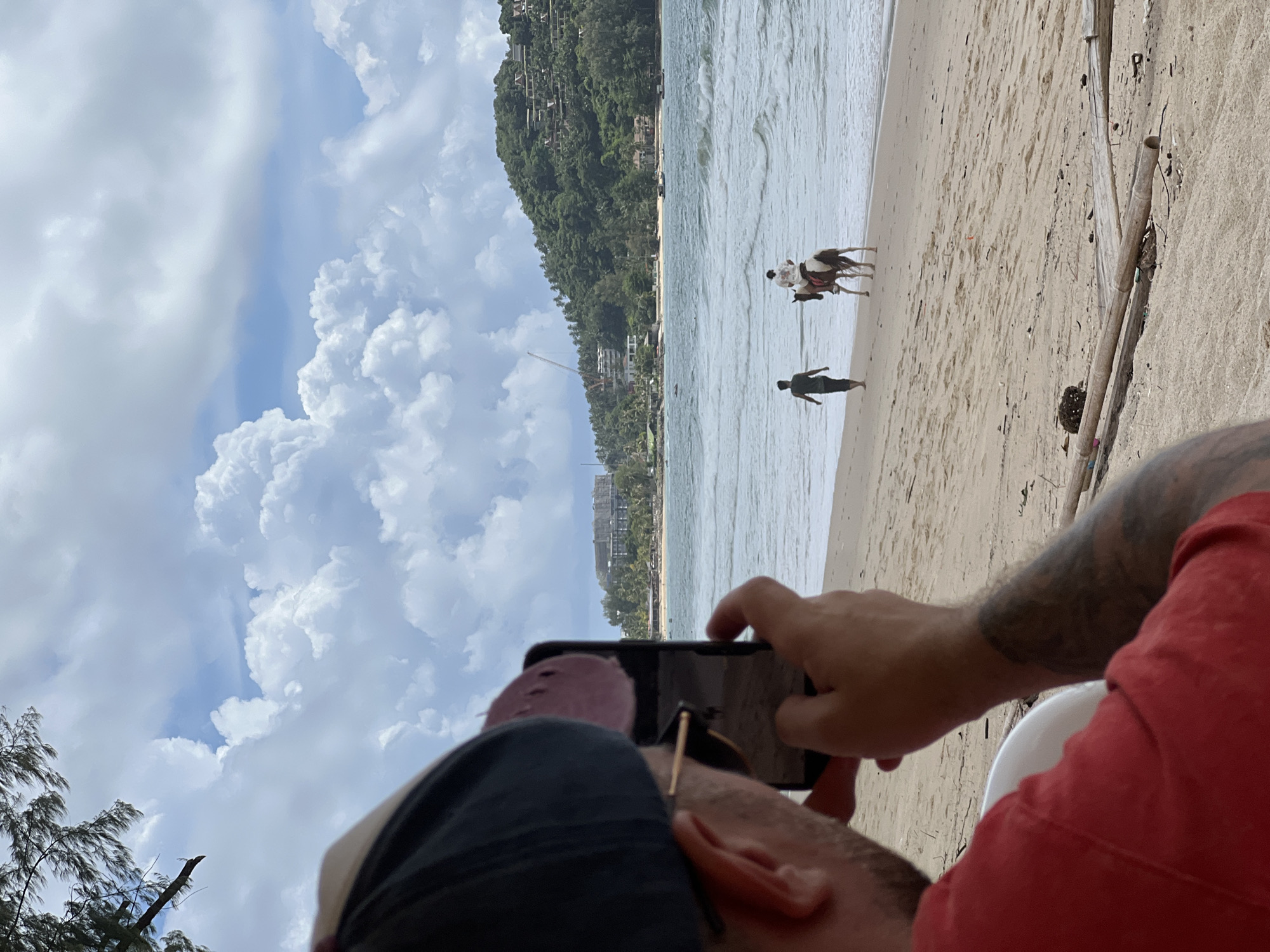 In Choeng Thale, Thailand, a person in a red shirt and a baseball cap is holding a smartphone, taking a picture of a beach scene. In the background, the ocean meets the sandy shore. Two people are on the beach: one walks towards the ocean, and another rides a horse. The landscape includes lush green hills with buildings, palm trees, and hotels. The sky is filled with large, white clouds. The foreground is dominated by the shoulder and arm of the person taking the photo, with visible tattoos on their arm. Beach debris, including bamboo sticks and dried leaves, is scattered on the sand.