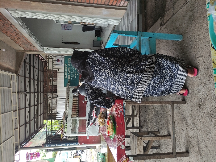 An outdoor, vertically oriented scene in Kepanjen, Indonesia, depicts a person seated at what appears to be a street vendor's stall during daytime. The individual, dressed in a long, dark blue or black patterned garment with white designs (likely a sarong or skirt) and pink flip-flops, is positioned on a low, rustic wooden bench. In front of them is a small, simple metal table or cart laden with various packaged food items, including what looks like instant noodles with the brand "INDOMIE" partially visible on a red banner. A dark scooter or motorcycle is parked nearby, partially seen to the left.

Behind the seated person is a light blue wooden bench. The background features several elements: a building with exposed red brick walls and a white upper facade, and a large open-air structure with a corrugated metal roof supported by metal trusses, suggesting a covered market area or a shaded walkway. A prominent green sign with white lettering is visible on a wall, clearly stating "BANTUAN HUKUM" (Legal Aid) and "BHIRAWAMA SATYA YUSTISI" below it. The ground is concrete, and the scene is brightly lit by natural sunlight, indicating clear weather. The overall setting suggests a casual, informal urban or semi-urban environment typical of Indonesian street life.
