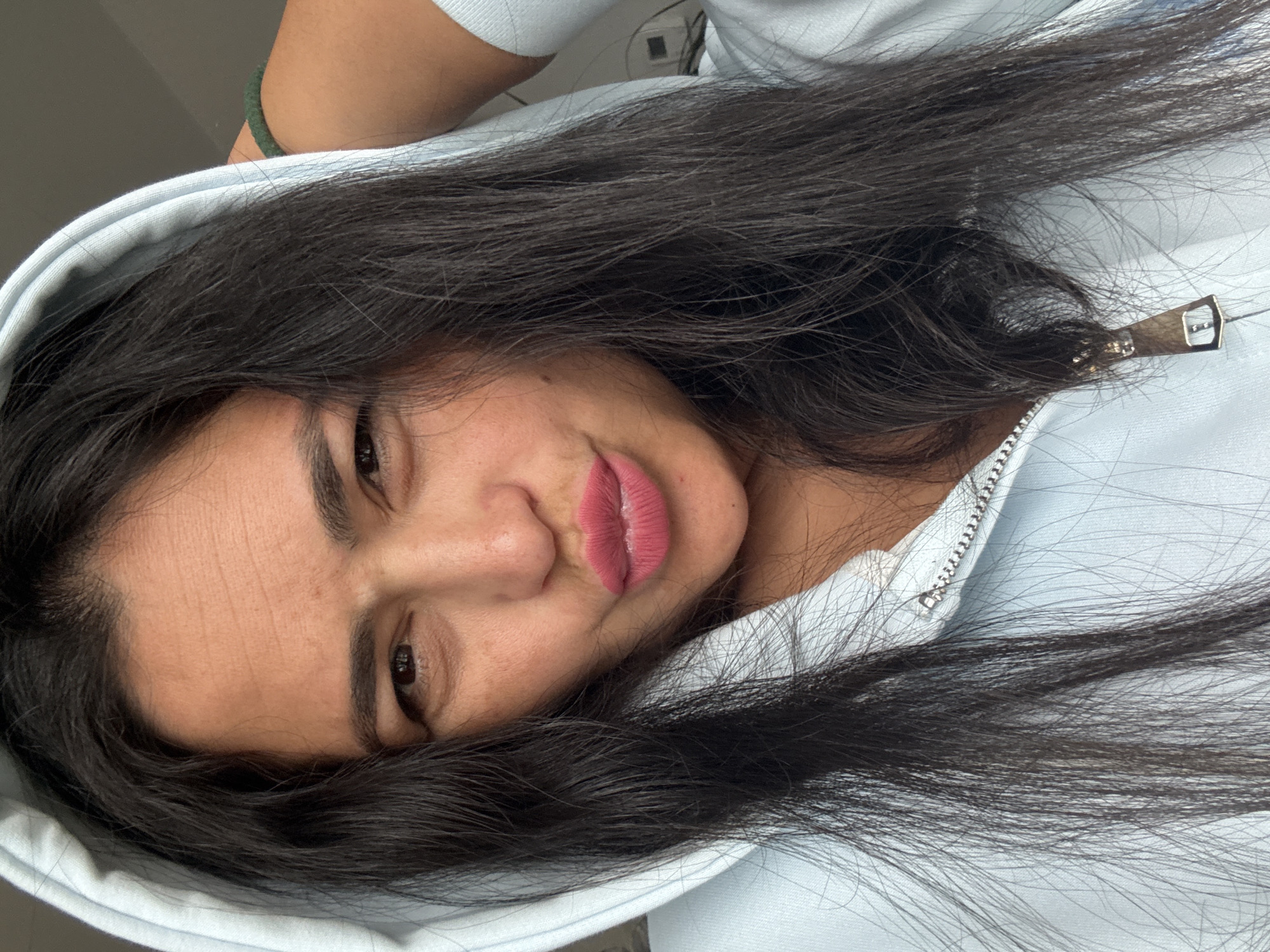 A woman with dark hair and light skin is lying down, wearing a light blue hooded sweatshirt. She is facing the camera with a slight smile. The scene is likely indoors and there are no other discernible people or objects in view to determine a location, including Kauchuk Stantsiya, Russia.
