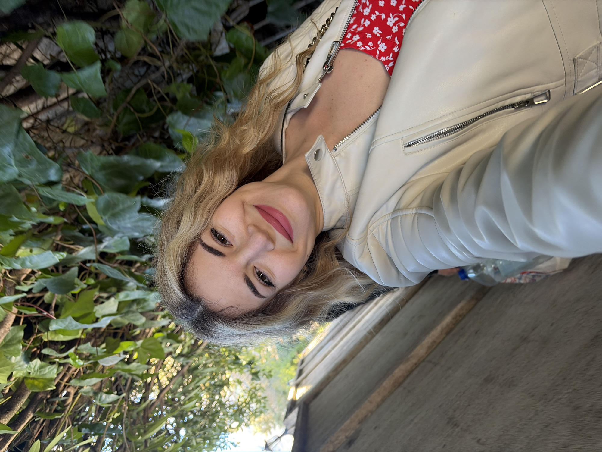 A close-up selfie photograph depicts a single individual, identified as female, outdoors during daylight hours. The subject, positioned centrally, has long, wavy blonde hair and a light skin tone. She exhibits a closed-mouth smile and has applied pink lipstick. She wears an unzipped, light beige/off-white zippered jacket with silver-toned hardware, revealing a red garment patterned with small white flowers underneath. Dense green foliage, consistent with ivy or similar climbing plants, occupies the upper and right portions of the background. A light brown, horizontally planked wooden surface occupies the lower-left background. A blue plastic bottle and a white bag are partially visible beneath this surface. Illumination is provided by natural daylight. The precise location within Budva, Montenegro, cannot be determined from the visual information presented.