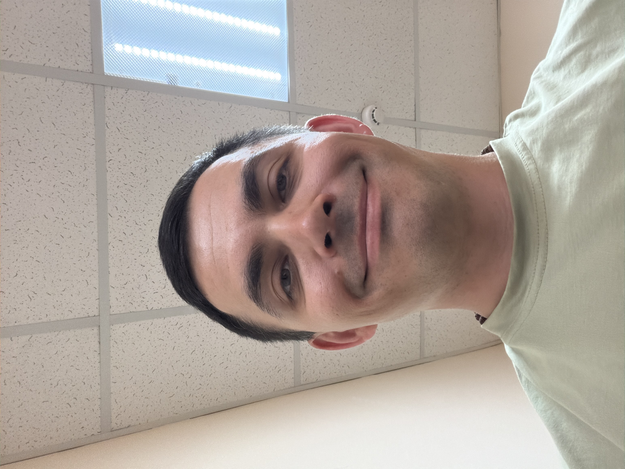 A man with short, dark hair is positioned with his face angled downward, looking into the camera. He has fair skin, dark eyebrows, and a slight smile. He is wearing a light green t-shirt. The background consists of a white acoustic ceiling with a fluorescent light fixture and a small circular object (likely a smoke detector). There is no indication of a location in the city of Izhevsk, Russia.