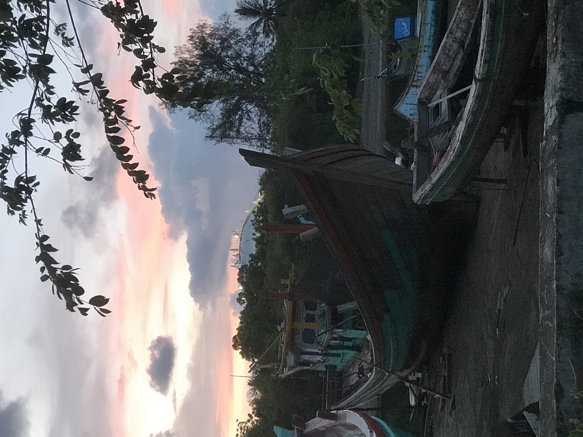 Several old, weathered fishing boats are on land, possibly for repair or storage. The boats are arranged in a line, with one boat appearing larger and more prominent than the others. The background features trees, a road, and a setting sun over the city of Lhokseumawe, Indonesia, as seen by the stadium lights near the shore. A tree branch with leaves is visible on the left side of the frame. A concrete wall is on the right side of the frame.