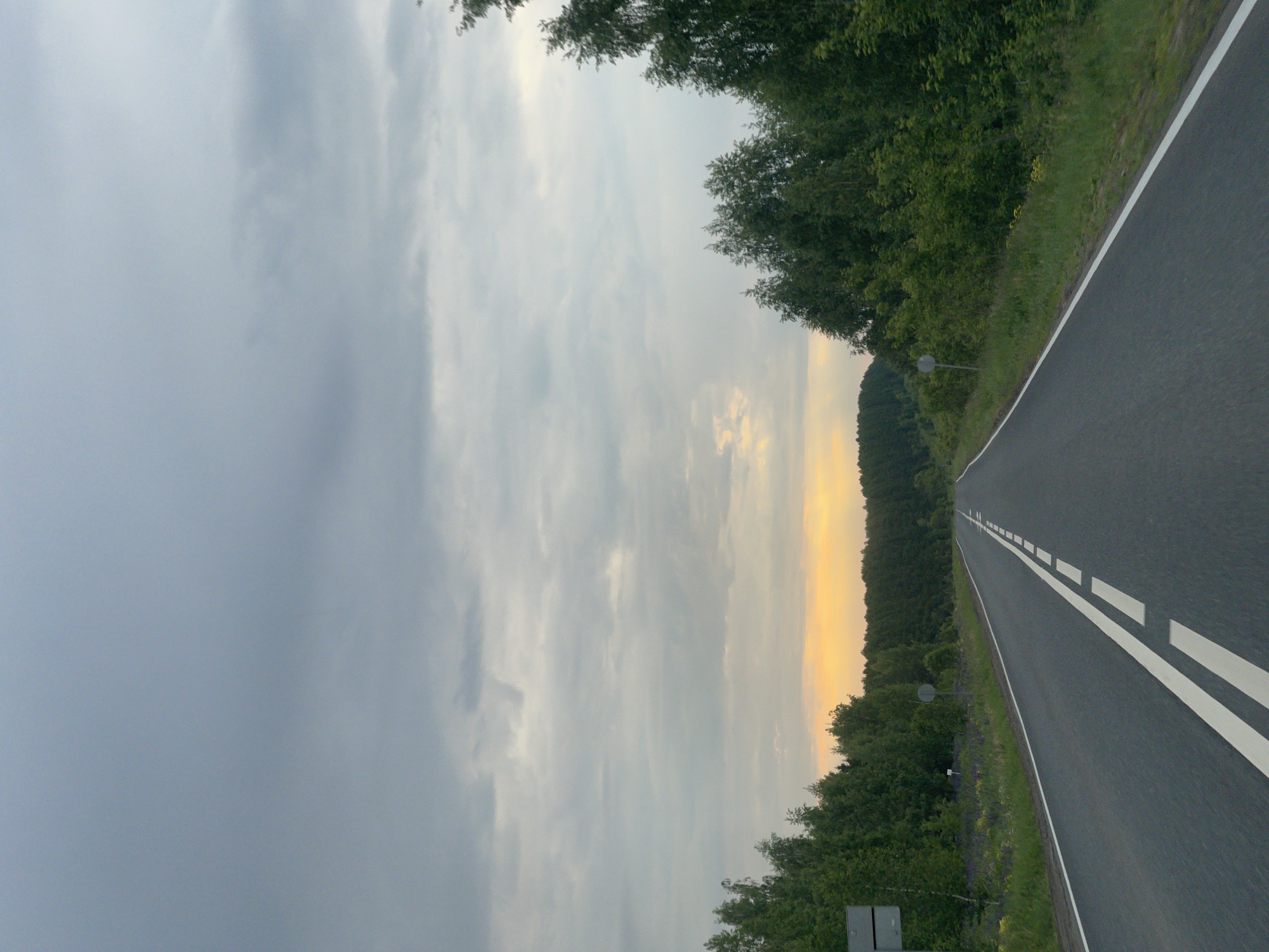 The image depicts a long, empty two-lane asphalt road stretching into the distance under a vast, cloudy sky. The road is marked with clear white lines: a double solid white line on the left side (from the viewer's perspective, given the likely orientation before rotation) and a dashed white line on the right, suggesting traffic lanes and passing zones. Both sides of the road are bordered by lush green grass shoulders and dense walls of green trees, forming a natural tunnel as the road recedes.
The sky is predominantly covered with light grey and white clouds, but a soft, warm orange and yellow glow is visible on the distant horizon, indicating either sunrise or sunset. This warm light contrasts with the cooler tones of the clouds and casts a tranquil atmosphere over the scene. A small, circular road sign is partially visible on the left side of the road, mostly obscured by the roadside foliage. There are no people or vehicles visible, giving the impression of solitude and open space. The overall mood is serene and expansive, capturing a moment of journey or quiet observation in nature. No discernible text is visible anywhere in the image.