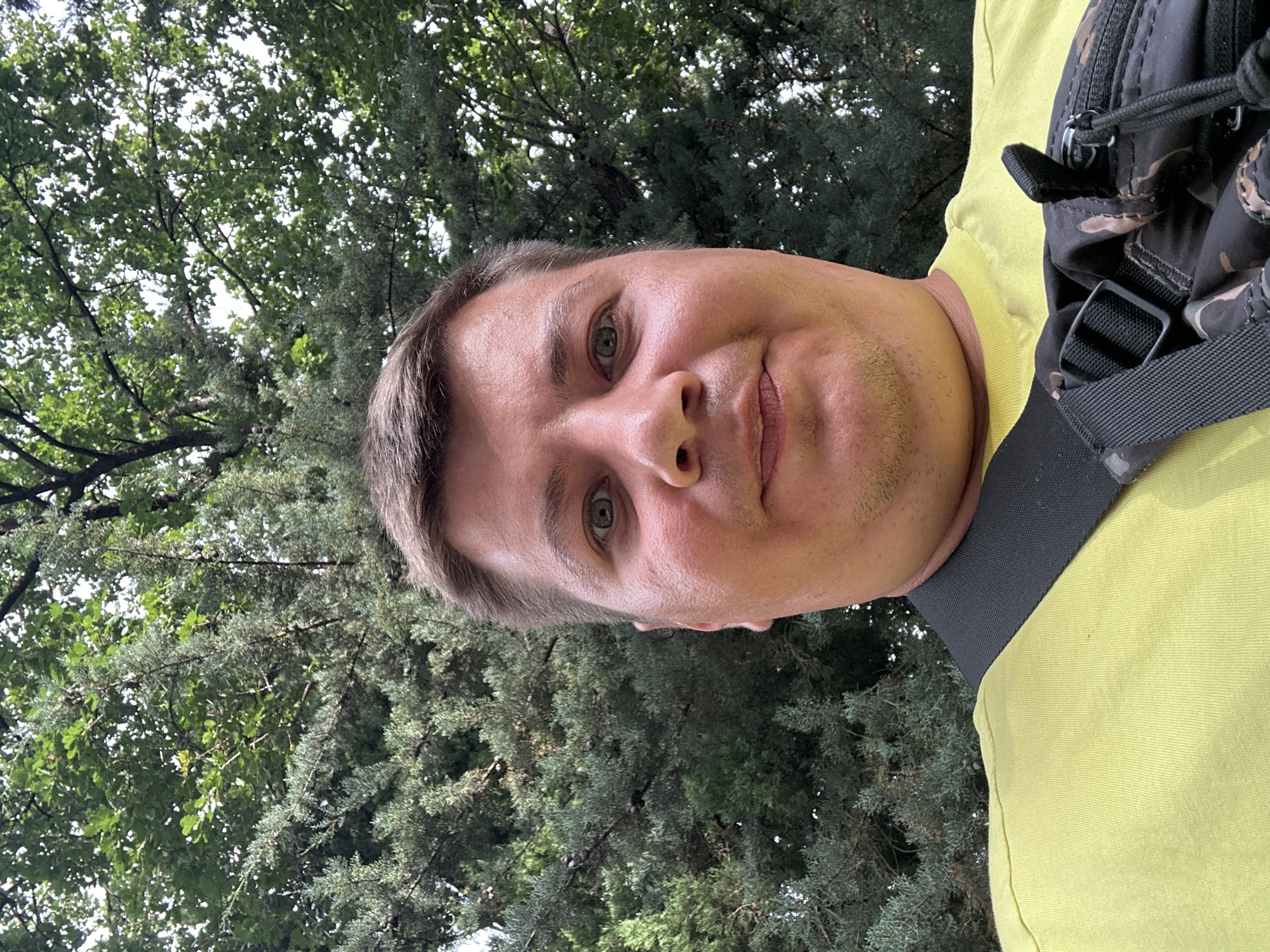 This is a selfie of a man with light brown hair and green eyes, looking directly at the camera with a slight smile. He is wearing a bright yellow t-shirt and a dark, patterned backpack is visible over his right shoulder. The photo appears to have been taken outdoors, with lush green trees and foliage forming the background. The lighting suggests it is daytime, likely with overcast or dappled sunlight filtering through the leaves. The setting could be a park or a wooded area, given the dense greenery. The orientation of the photo is vertical, as if taken with a phone held upright. The man's expression is casual and relaxed. No text is visible in the image. The location context provided is Budva, Montenegro.
