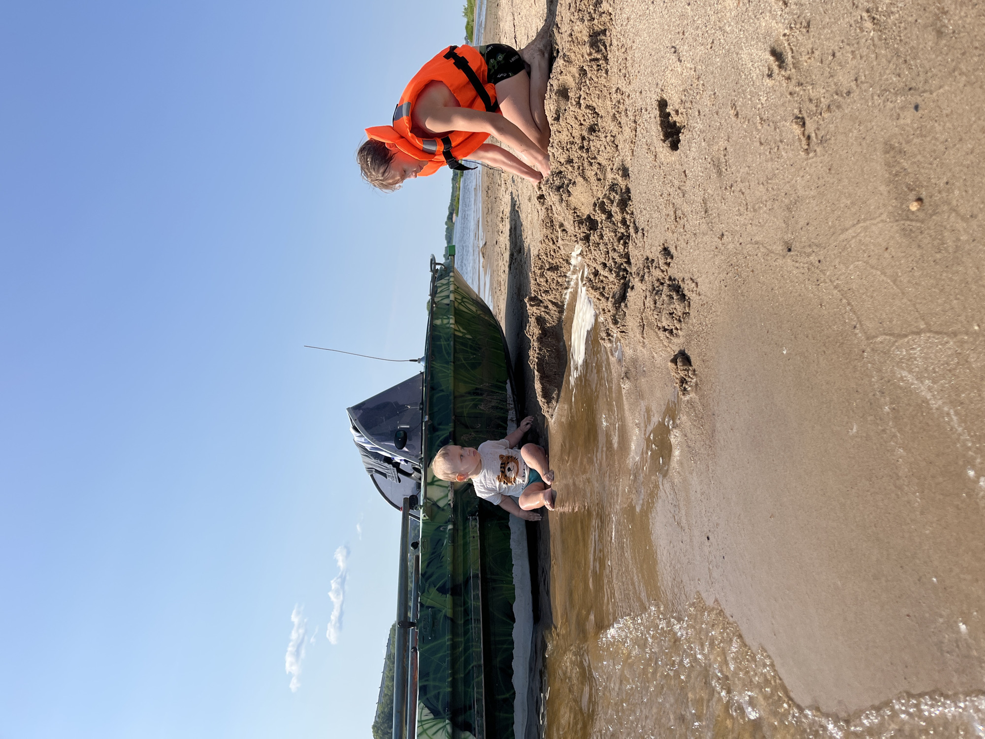 The image depicts a sunny day at the beach, likely on the banks of the Volga River in Nizhny Novgorod, Russia. A small, camouflage-patterned boat is partially submerged in shallow water near the sandy shore. To the left, a toddler sits in the water's edge, facing away from the camera, wearing a light-colored t-shirt and blue swim trunks. To the right, a young person, wearing an orange life jacket and black shorts, is kneeling on the sand and appears to be playing with the sand. The sky is clear blue with a few wispy clouds. The water is calm, and gentle waves are lapping at the shore. The overall scene is one of outdoor recreation and relaxation.