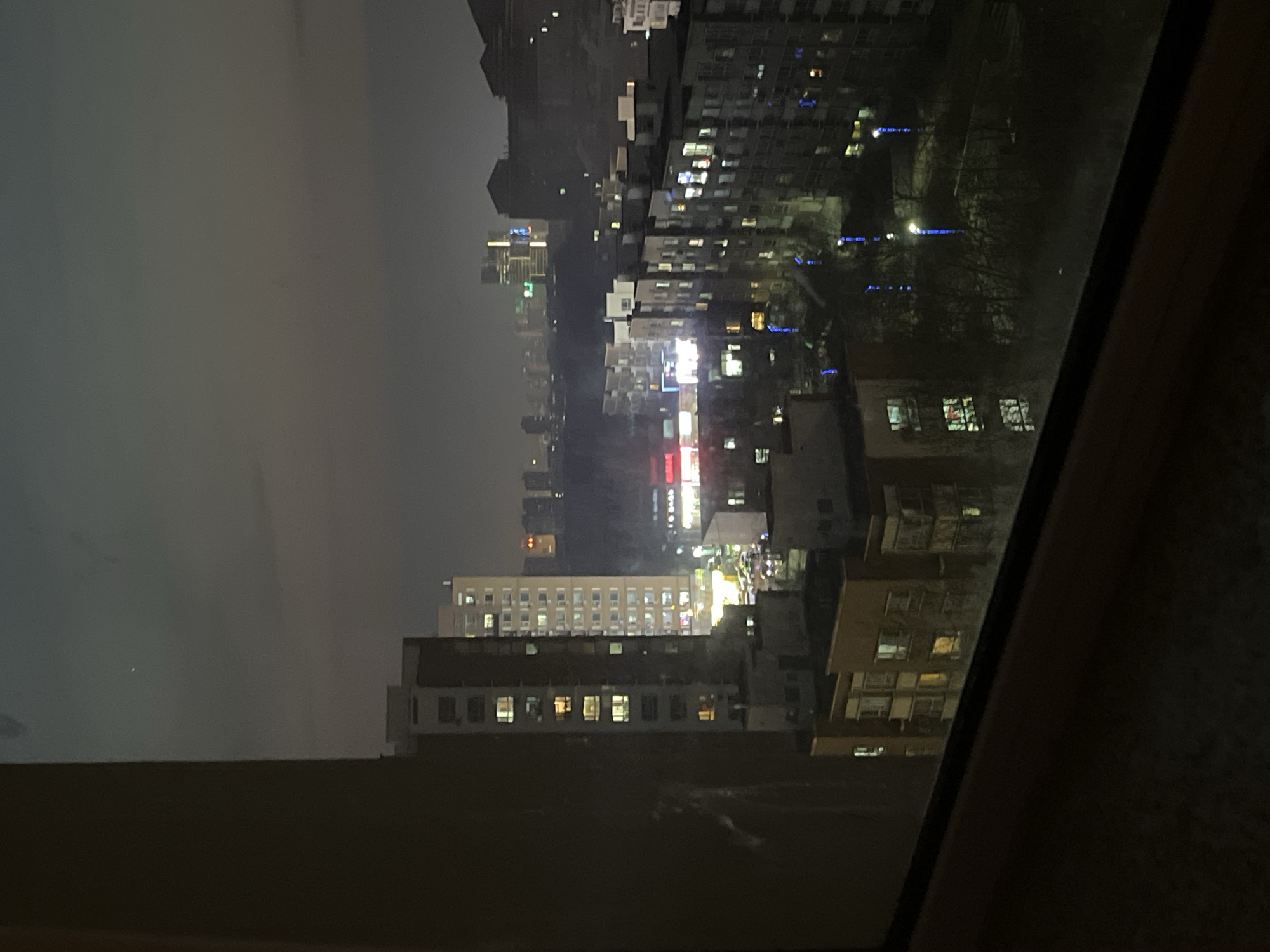 The image shows a night view of Changchun City, China, taken through a window. Several buildings are visible, lit with various lights. In the foreground, parts of the window frame are visible, suggesting the photo was taken indoors. The sky is dark, and atmospheric conditions seem foggy or hazy, muting distant city lights.