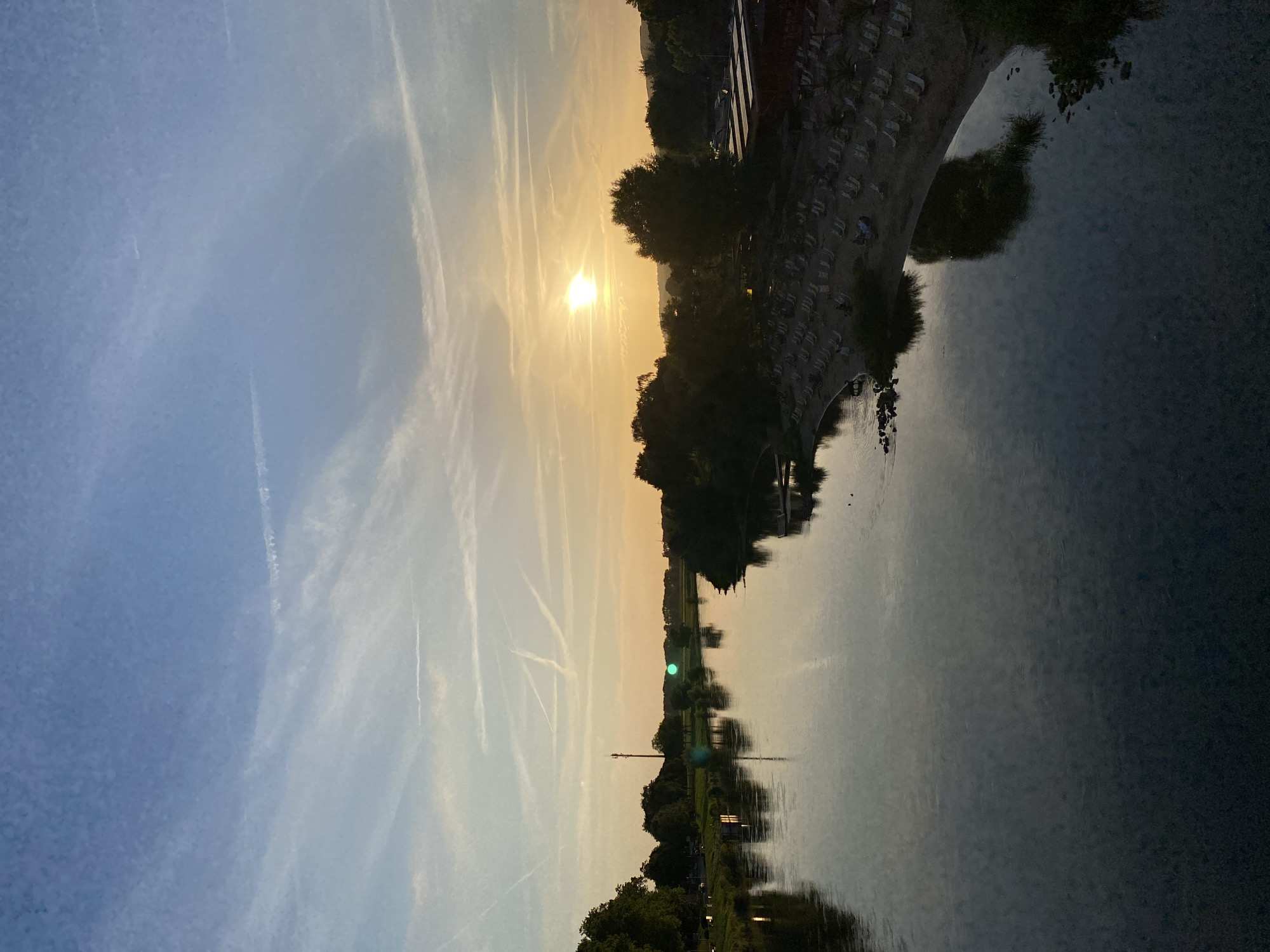 A wide river flows through the city of Rinteln, Germany, at sunset. The sky above is a gradient of blue and yellow, with wispy clouds streaked with condensation trails. The sun, a bright orb, is setting on the horizon, casting a warm glow across the water and the landscape. The river's surface is calm and reflects the colors of the sky and the surrounding trees. On the right bank, a sandy beach is lined with numerous empty lounge chairs, suggesting a leisure area. Dense green trees and foliage line the riverbanks on both sides. In the distance, a tall tower with an antenna stands on the left bank, and a low-rise building with a red roof is visible on the right bank, near the beach. The overall atmosphere is serene and tranquil.