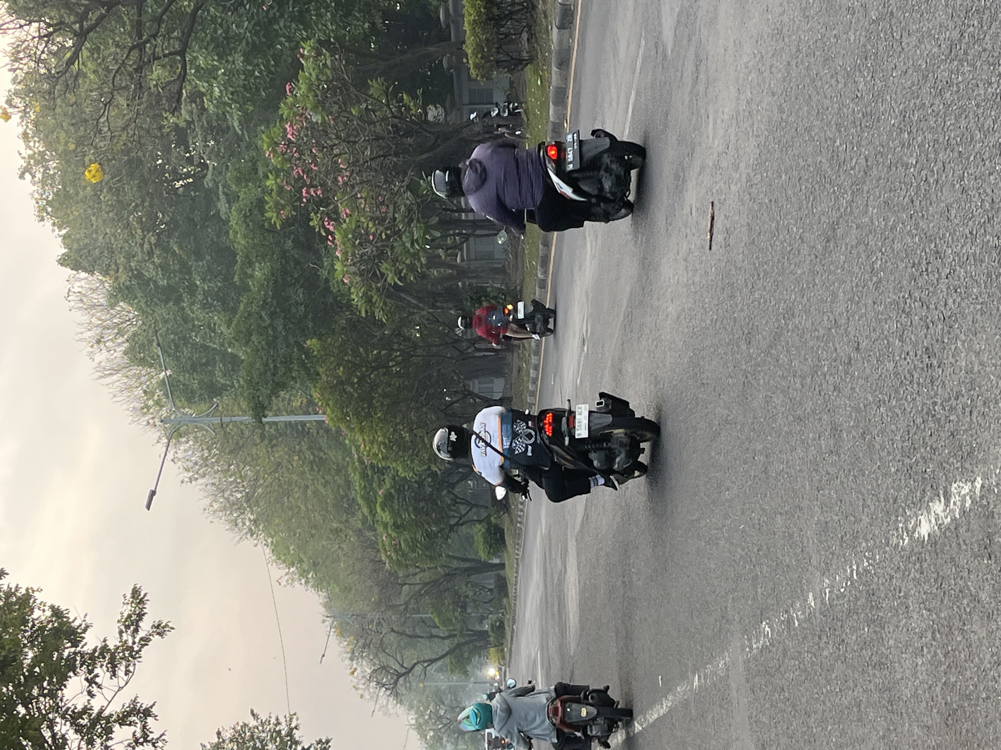 The image depicts a street scene in Gelam, Indonesia. Several individuals are riding motorcycles on a paved road lined with trees and vegetation. The sky is overcast, suggesting it might be early morning or late afternoon.

In the foreground, a person in a blue helmet and gray hoodie rides a scooter from left to right. To their right, another person wearing a white shirt with a graphic design and a black helmet rides a scooter. Further ahead on the right side of the road, two more individuals on scooters are visible; one in a red jacket and another in a purple hoodie.

The road has visible white lane markings. Streetlights are present along the side of the road. The environment appears to be a well-maintained urban or suburban area with ample greenery.