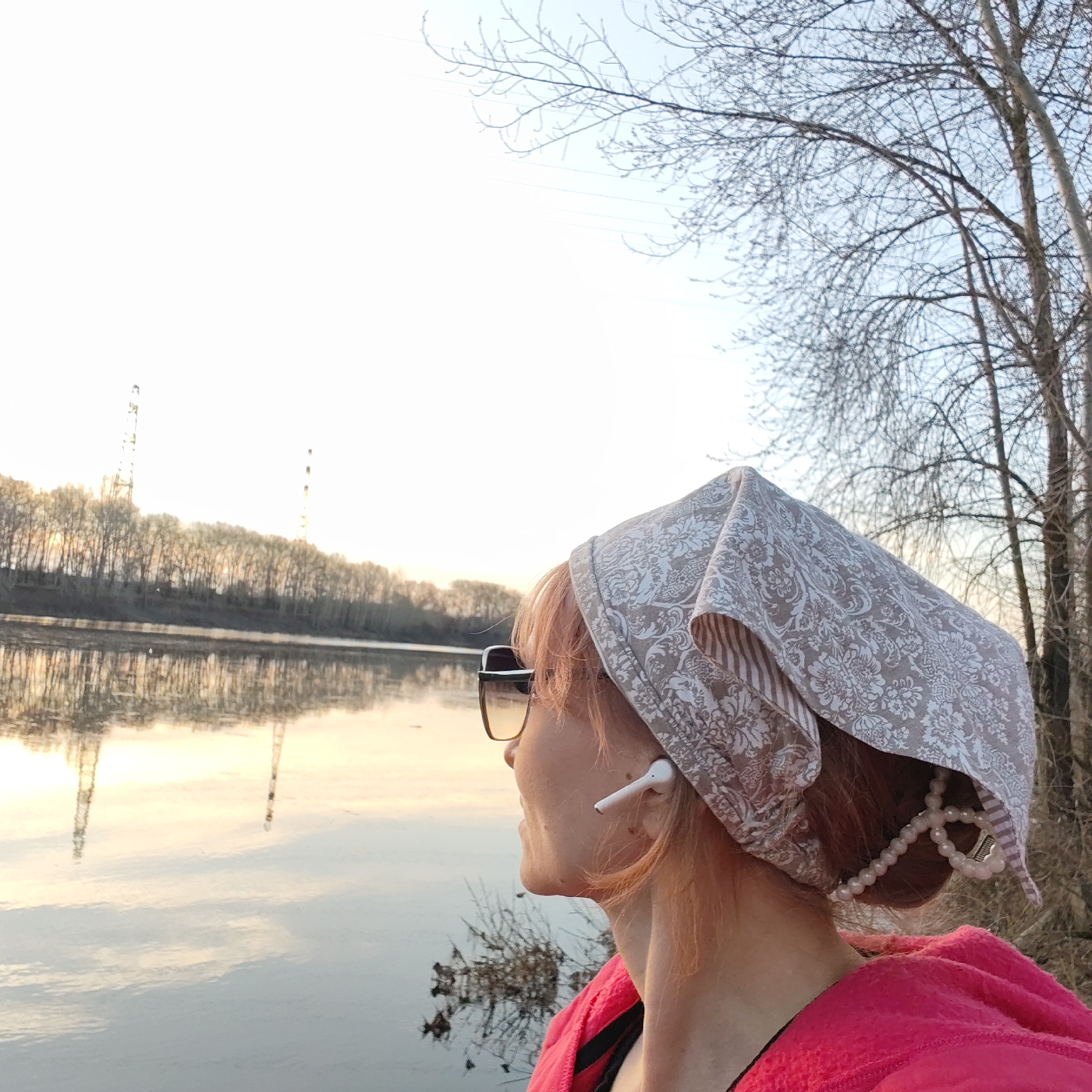 A woman wearing sunglasses and a patterned headscarf is looking toward a river with trees lining the opposite bank and two transmission towers visible in the distance. Her face is in profile. The river reflects the sunset and the towers. The trees in the background are bare, suggesting it's early spring or late autumn. The location is Chaikovsky, Russia. The woman has an earpod in her ear. A string of pearls is visible underneath the headscarf. The woman is wearing a pink fleece jacket.