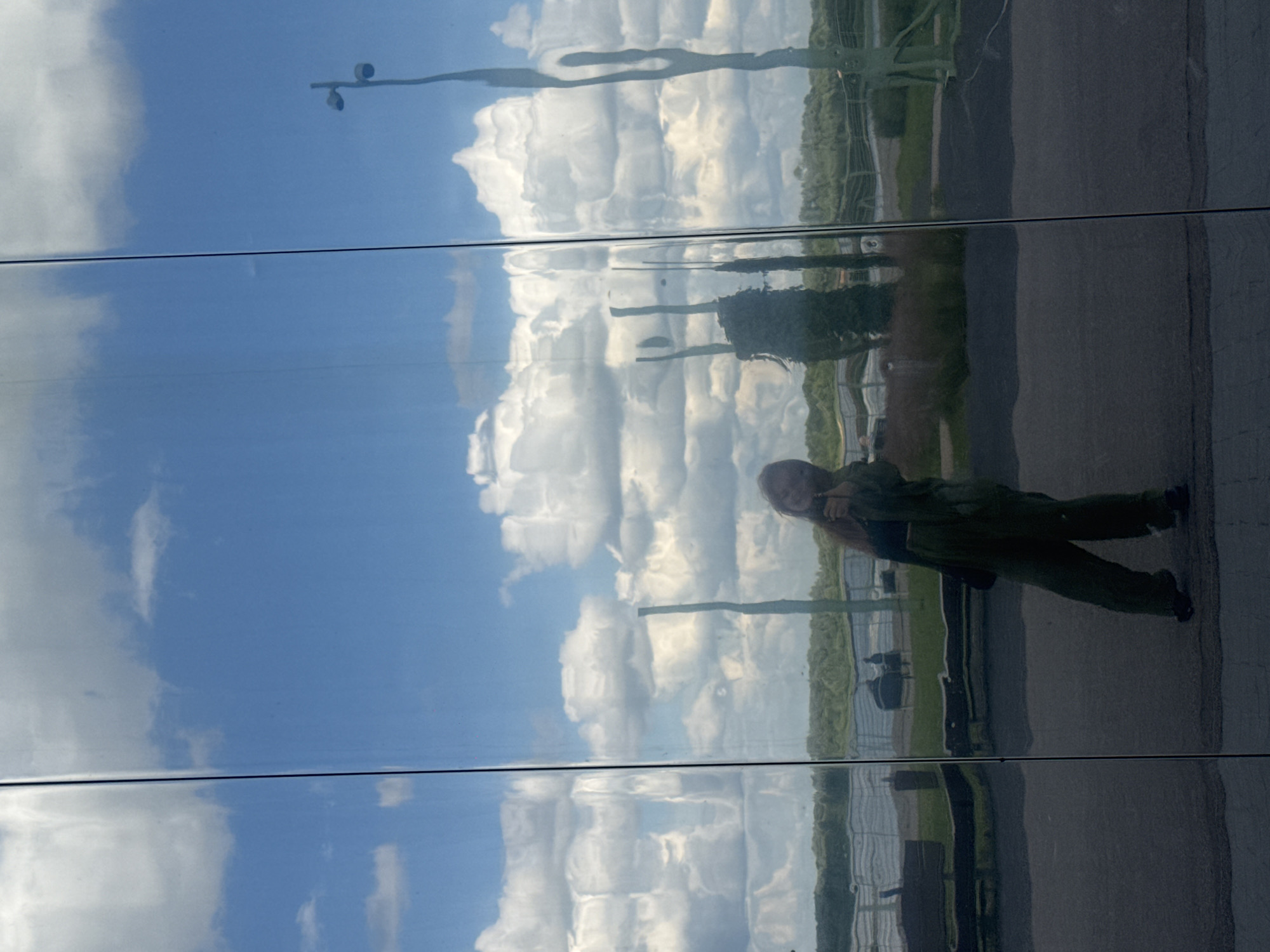 The image displays a reflection on a mirrored building surface. In the reflection, a young woman is standing and taking a selfie. She is wearing a green jumpsuit and holding her phone up to take the picture. Behind her, the reflection shows a bright blue sky with scattered white clouds, a line of green trees, and some outdoor seating areas. The mirrored surface distorts the reflection in places. The woman is positioned in the lower right quadrant of the image. The scene is set outdoors on a sunny day. This image does not provide any information about the city of Nizhny Novgorod, Russia.