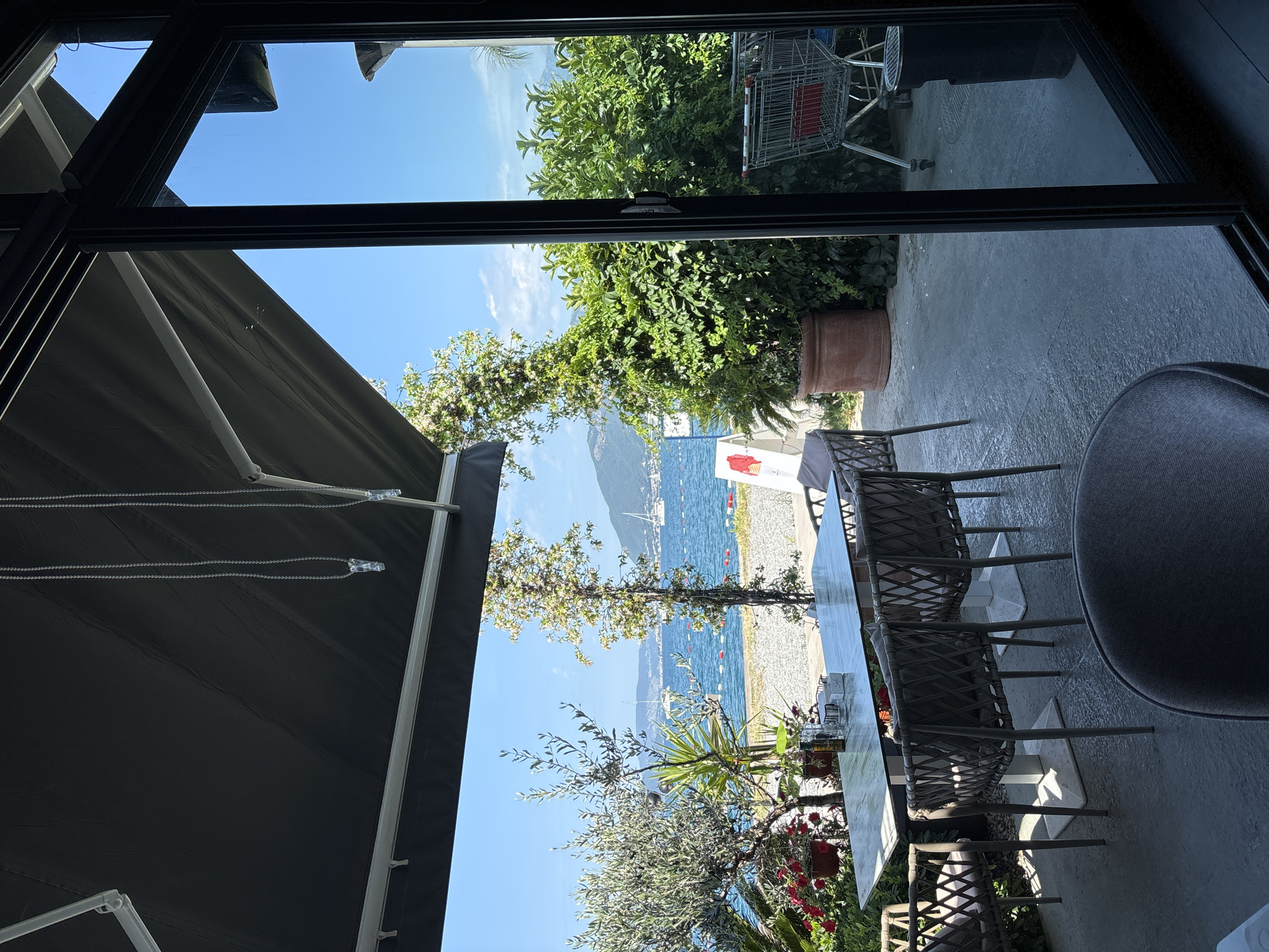 View from an open doorway of an outdoor dining area in Tivat, Montenegro. The scene captures a table and chairs arranged on a patio, with a partial view of a seated person. Outside, there is a view of a beach, trees, and a body of water with boats. A mountain is visible in the distance. A retracted awning partially covers the view from the left, and greenery is present in planters and surrounding areas. A shopping cart is seen near a building exterior behind the planter. The sky is clear and blue.